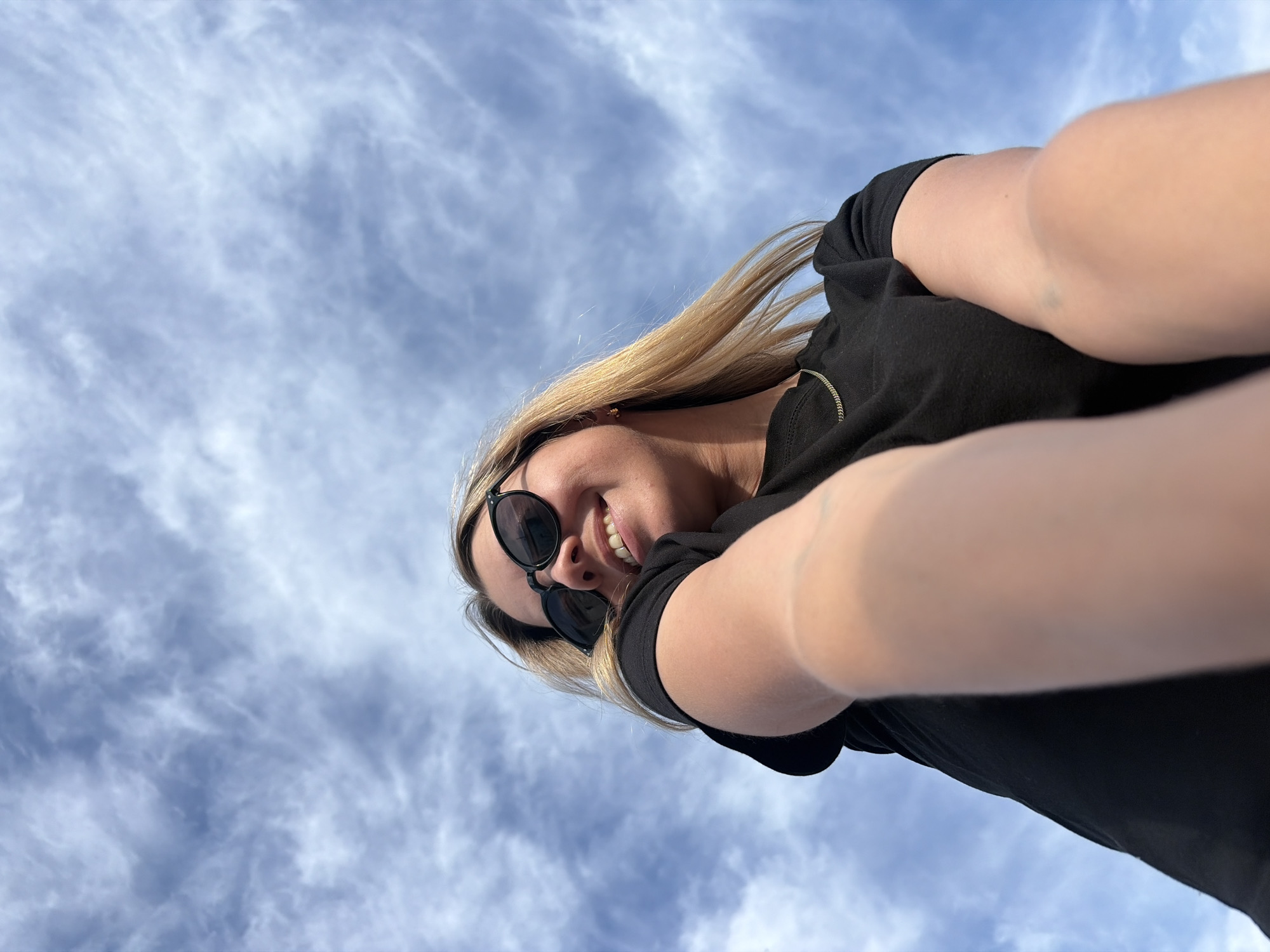 A woman is taking a selfie outdoors. She has blonde hair and is wearing a black shirt and sunglasses. She is smiling and looking at the camera. The background is a cloudy blue sky. The image appears to be taken during the day, and the weather looks pleasant. The woman's arms are extended towards the camera, as if she is holding a phone or camera to take the picture. There are no visible text elements in the image. The location context provided is Chaikovsky, Russia, but there are no specific visual cues in the image to confirm this location.