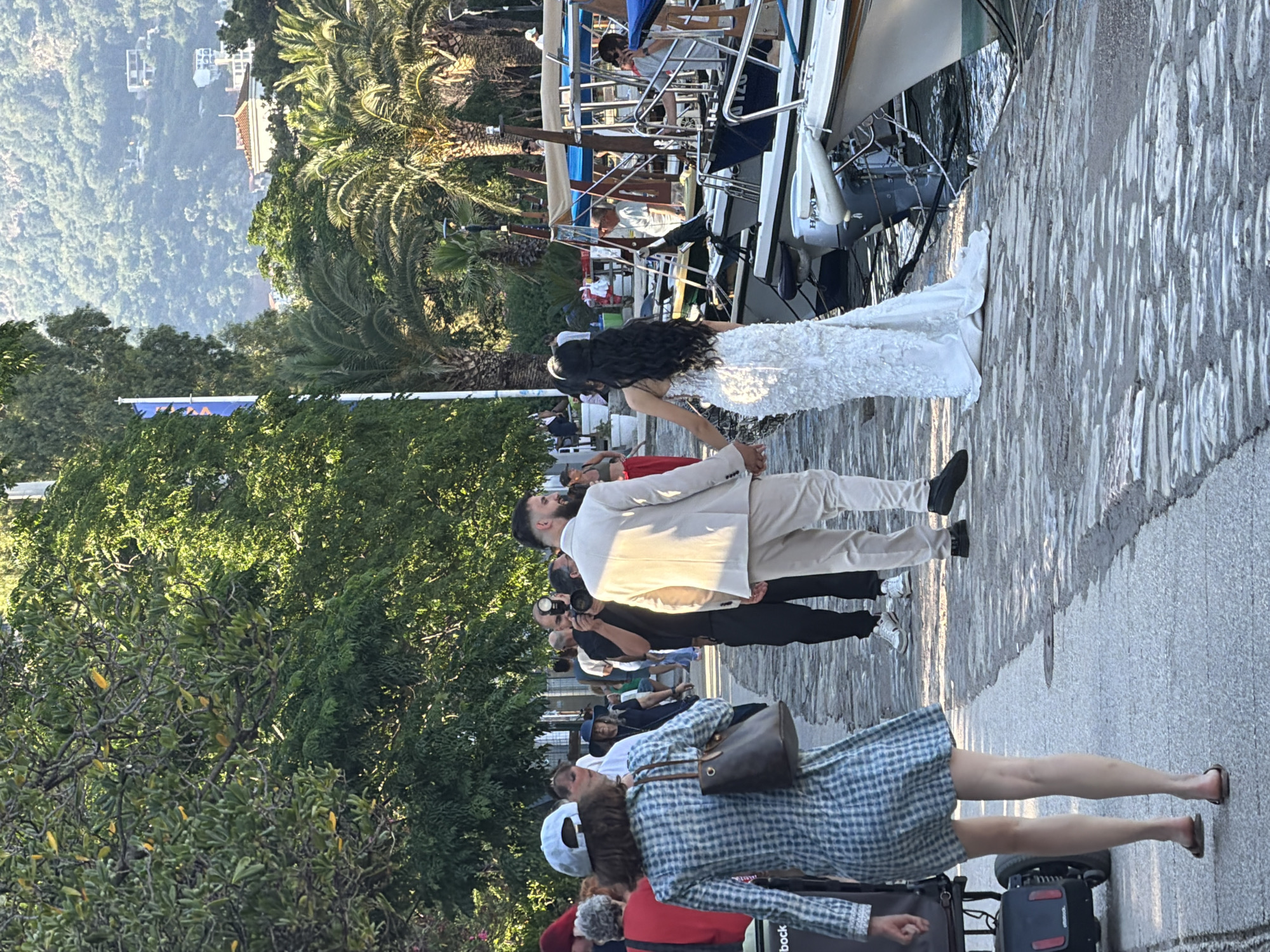 A wedding party is walking along a waterfront promenade in Budva, Montenegro. A bride in a white lace dress and a groom in a light-colored suit are holding hands and walking away from the camera. They are followed by a photographer who is taking their picture. Other people, some dressed casually and others more formally, are also walking along the promenade. Boats are docked along the right side of the image, and a hillside covered with trees and buildings is visible in the background. The promenade is paved with cobblestones, and trees line the left side of the path. The scene appears to be taking place in the late afternoon, with warm sunlight casting long shadows.