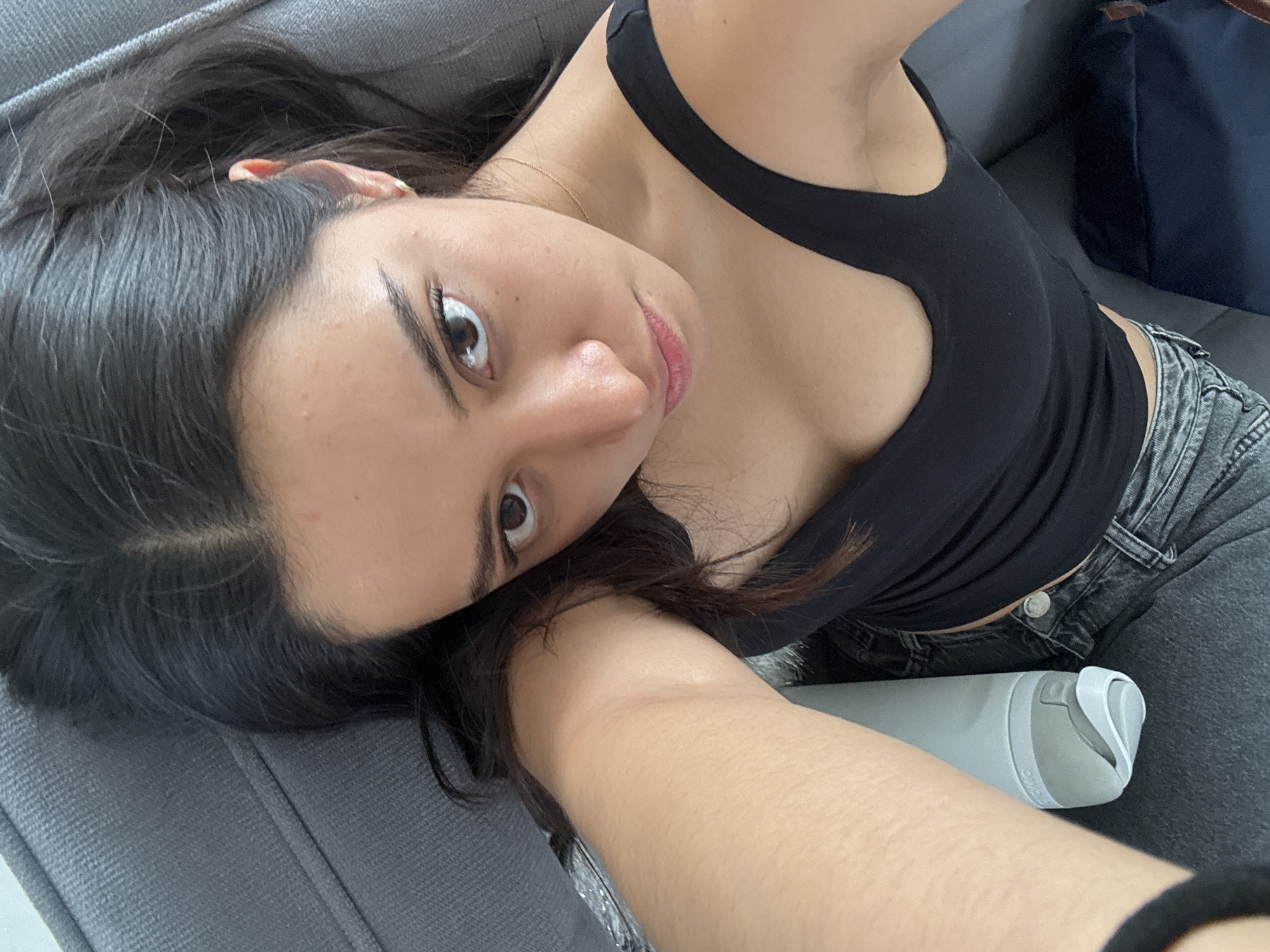 A medium shot shows a light-skinned woman with dark hair and eyes lying on a grey couch, facing the camera. She is wearing a black tank top and grey jeans. A grey water bottle is partially visible near her waist. A navy bag sits on the couch behind her. The location is Puebla, Mexico.