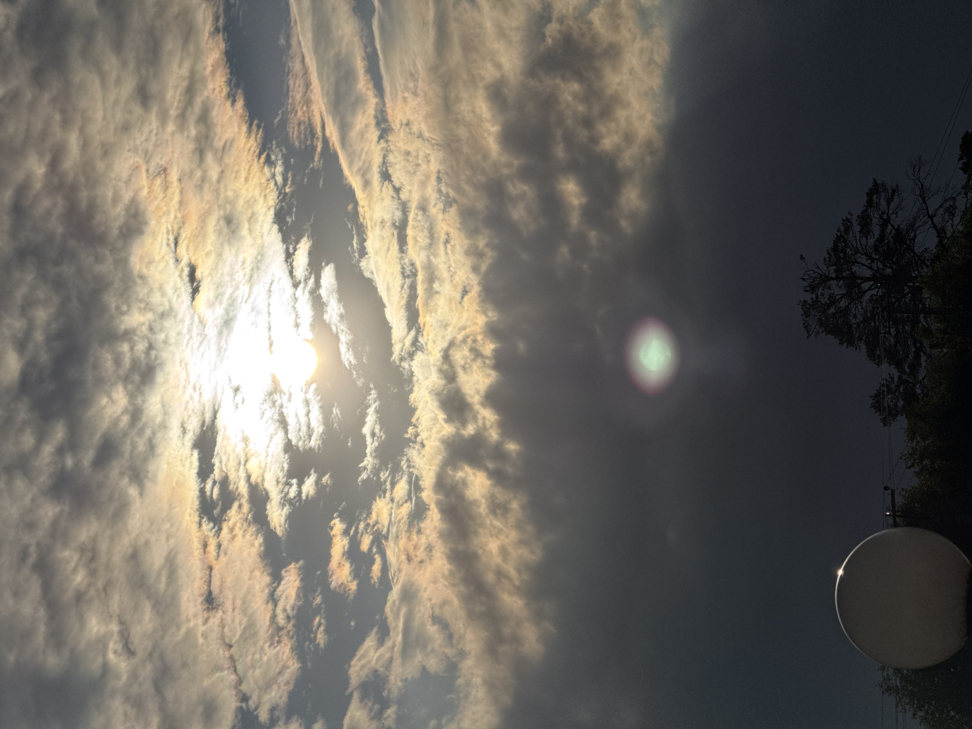 This media file depicts a view of the sky at dusk in Votkinsk, Russia. The sun is partially obscured by clouds, casting a warm, golden light on some of them, while others appear dark and heavy. The sun itself is a bright, blown-out white disc. There is a visible lens flare in the upper right quadrant of the image, and another fainter one towards the lower left. In the background, silhouetted against the dimming sky, are the bare branches of a tree and what appears to be a satellite dish, illuminated by a faint light source. The overall mood is somewhat dramatic due to the contrast between the bright sunlit clouds and the dark sky. The image is oriented vertically. There is no visible text.