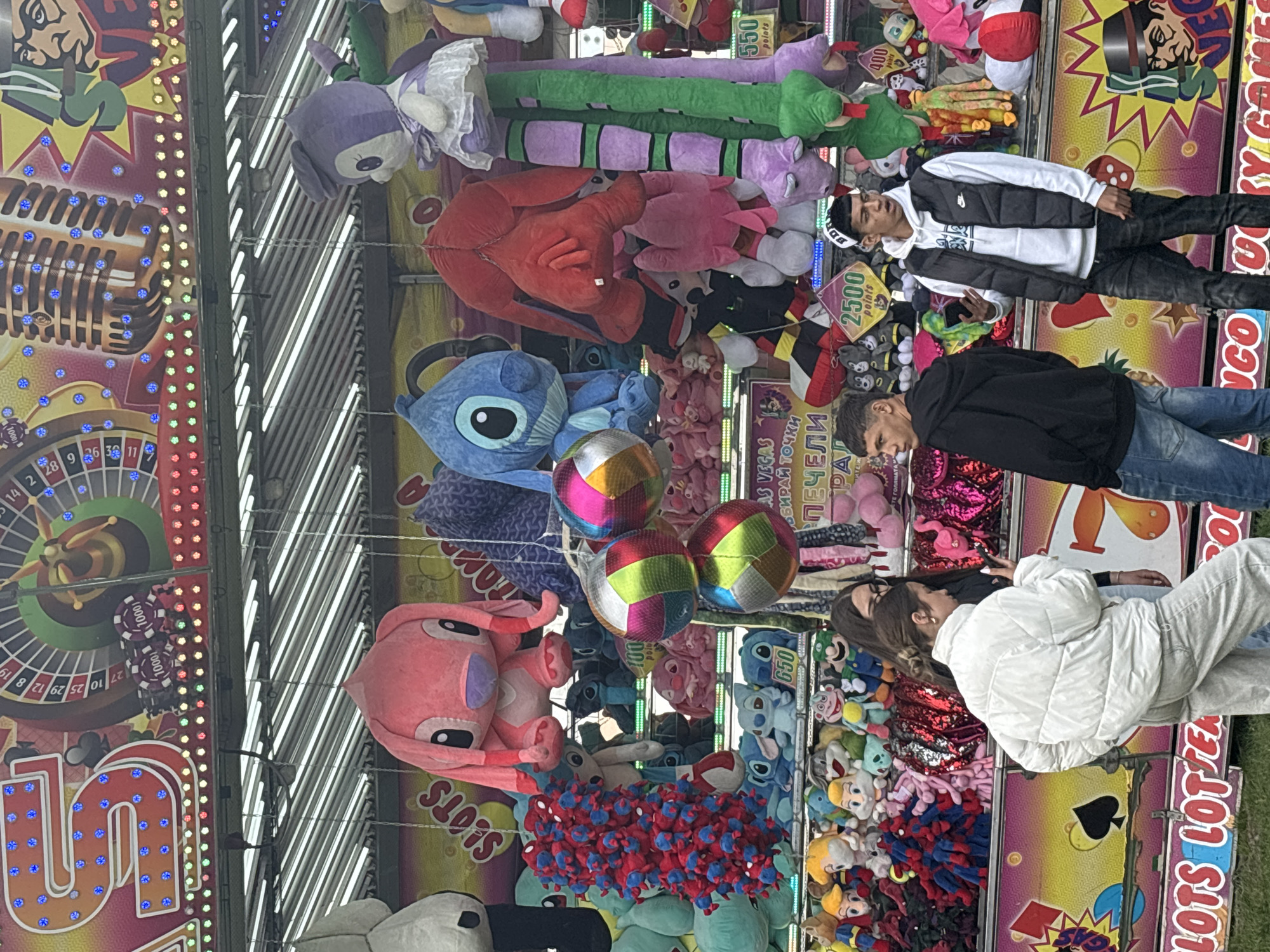 Here is a detailed description of the image:
**General Overview:**
The image depicts a carnival-like setting with various plush toys displayed as prizes in a game. People are standing near the booth, presumably interested in playing. The scene is colorful and well-lit.
**People:**
- There are people visible, some of whom appear to be interacting with the game booth.
- There are two men standing to the left side of the image. One is wearing a black and white vest, black shirt, and black pants, and the other is wearing a black jacket and blue jeans.
- There are two women standing to the right side of the image, one wearing a puffy white jacket.
**Objects:**
- Various plush toys including animals and characters are hung or displayed.
- Several hanging colorful striped balls.
- Game booth signage with text in both English and another language.
- Game graphics with a roulette wheel.
**Activities:**
- People are potentially engaging in or observing a game where they can win prizes.
**Location:**
Although the image cannot confirm this, text on the booth suggests the location might be "AS VEGAS" and the city of Sofia, Bulgaria.
**Environment:**
The environment is characterized by bright colors, game graphics, and the presence of recreational activities that are indicative of a carnival or fair.