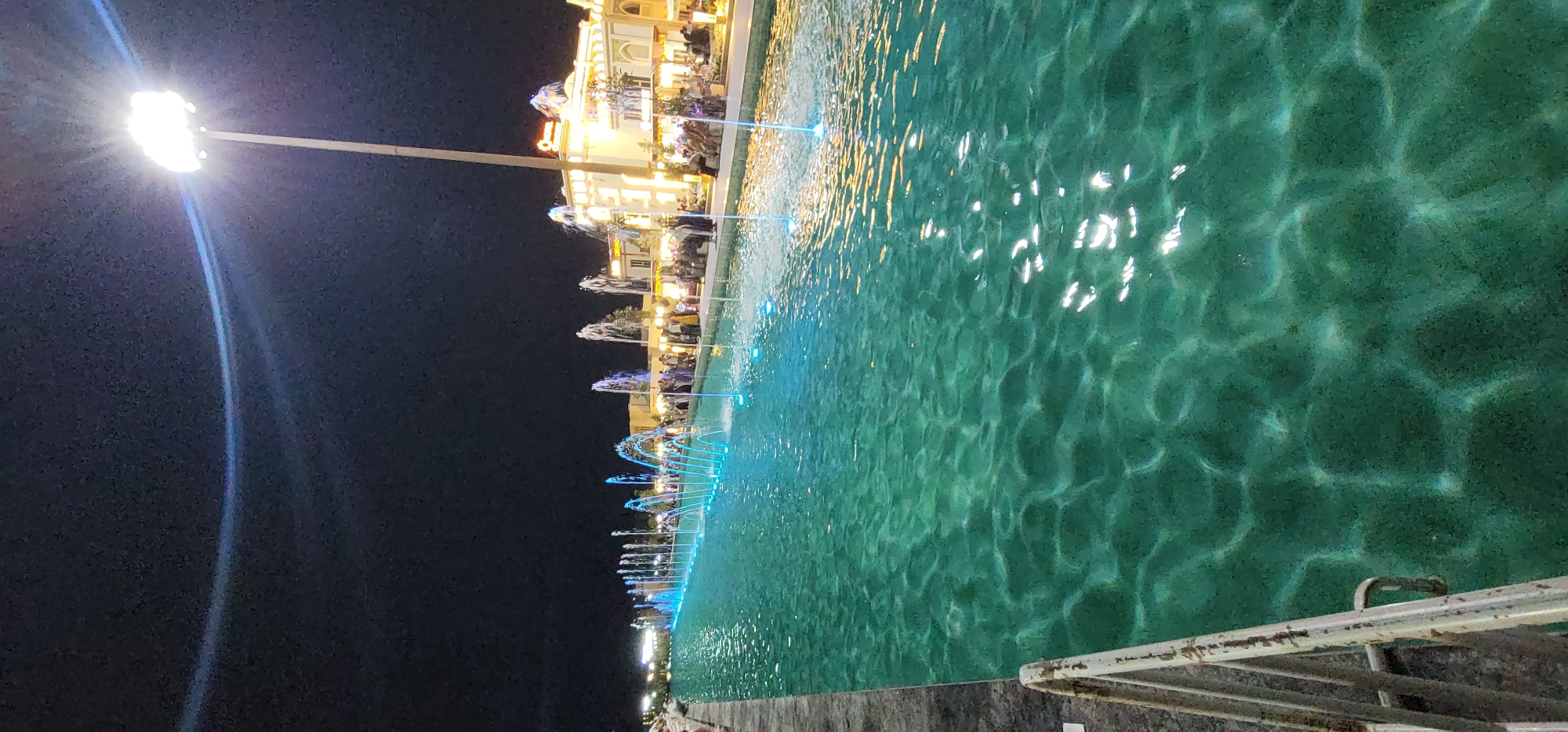 This image depicts an outdoor scene at night, likely in the city of Satkak, Uzbekistan, featuring a long, narrow canal or waterway lined with fountains. The water is illuminated with blue and white lights, creating a vibrant and dynamic effect. Along the far side of the waterway, a building with ornate architecture and illuminated windows is visible, suggesting it might be a commercial establishment or a public building. A bright floodlight on a tall pole is positioned on the right side of the frame, casting light over the scene and illuminating the night sky. Several people are gathered along the edge of the waterway, some appearing to be seated and others standing, suggesting a public gathering or recreational area. The overall atmosphere is one of nighttime entertainment and public space utilization.