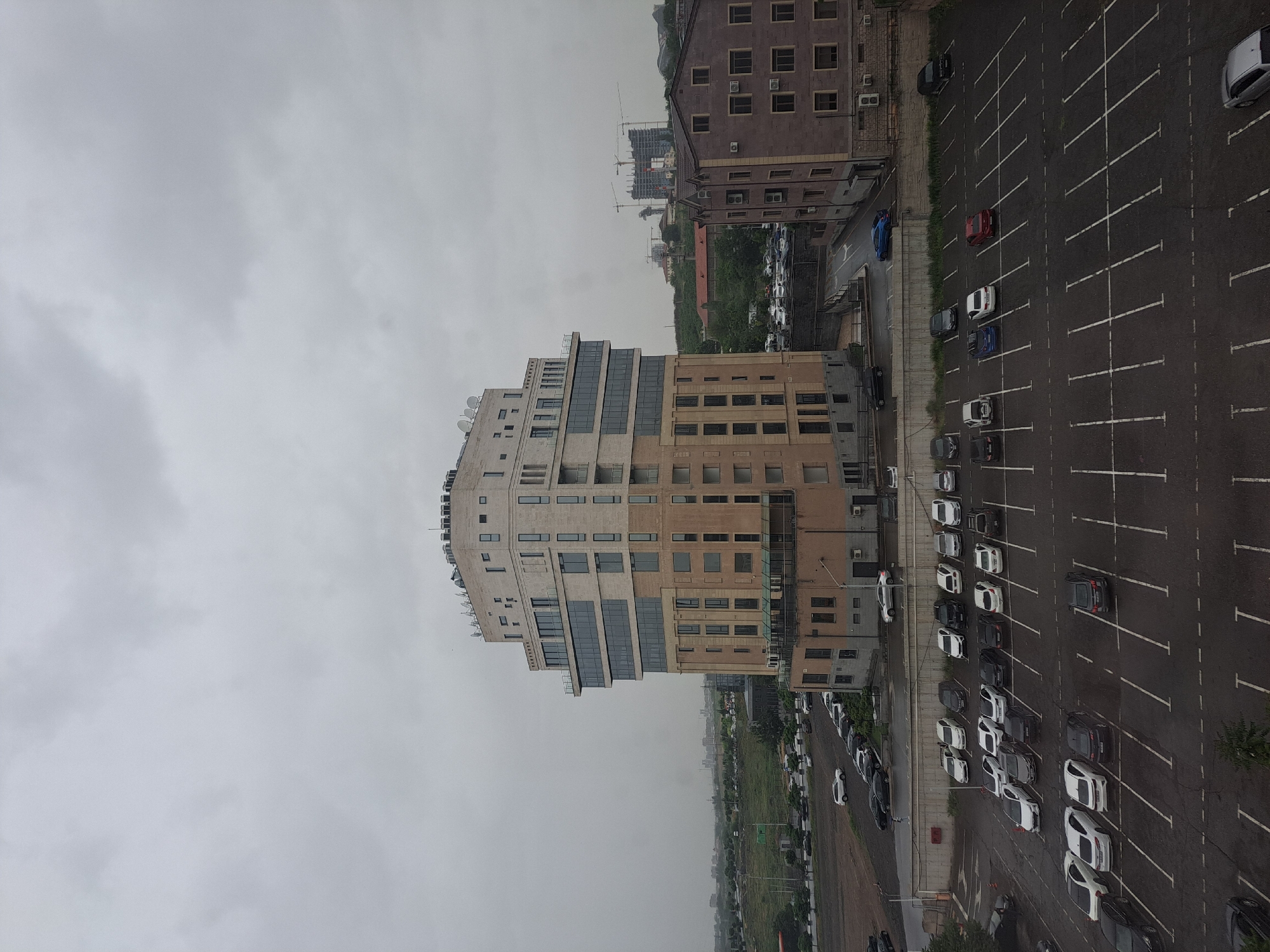 The image depicts a cityscape in Yerevan, Armenia, under an overcast sky. Dominating the scene is a modern multi-story building constructed with a mix of light and dark stone or concrete. The building's architecture is characterized by stepped setbacks, horizontal window bands, and satellite dishes on its roof. Adjacent to this structure is a large parking lot filled with various cars, marked with white parking lines. A second, darker-toned building with a more traditional design stands beyond the parking lot, featuring regularly spaced windows and visible air conditioning units. In the background, glimpses of other buildings, greenery, and construction cranes suggest urban development. The overall impression is one of a densely populated urban area undergoing continued growth and modernization.