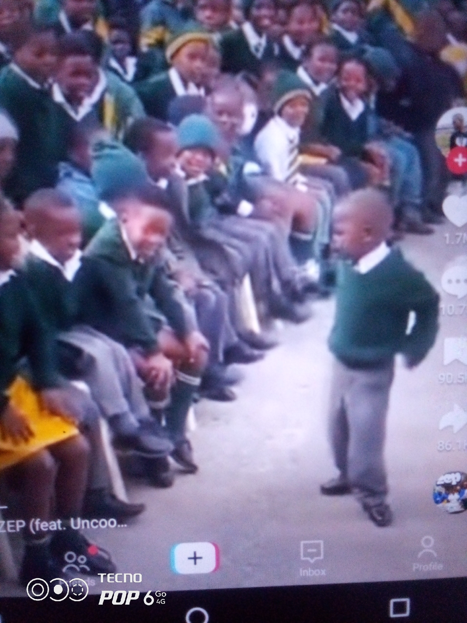 The image shows a group of young children, likely students, sitting and standing outdoors. They are wearing school uniforms consisting of green sweaters or blazers over white shirts, and grey pants. Some of the children are wearing green caps. The children appear to be in a schoolyard or an open area, possibly an assembly or a break. The background is blurry but suggests an outdoor environment with some structures. The image appears to be a screenshot from a mobile device, as indicated by the overlay of icons and text, including "ZEP (feat. Uncoo...", "TECNO", and "POP 6 4G". The presence of a red heart icon and a speech bubble icon with numbers next to them suggests a social media interface. There is no specific information in the image that links it to the city of Howayi, Nigeria.