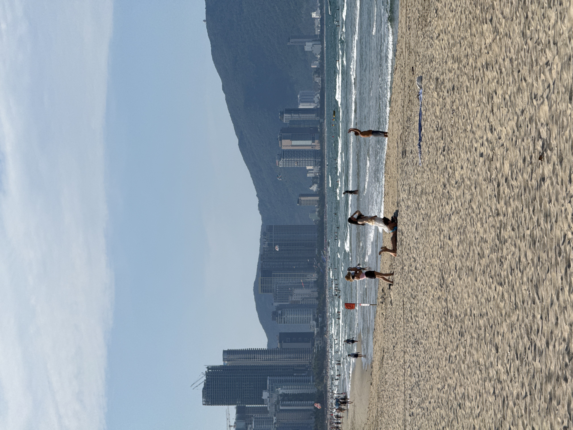The image depicts a beach in Đà Nẵng, Vietnam. A mountain ridge and numerous high-rise buildings line the horizon. The beach is sandy and populated with people. There are people swimming, standing, and laying on the beach. The waves are moderate. A red flag is planted in the sand near the water.