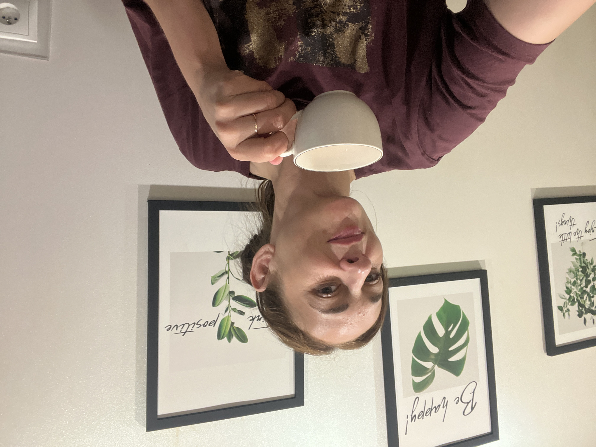 An image shows a woman with medium brown hair pulled back in a bun, holding a white teacup with a gold rim in her right hand. She is wearing a dark maroon t-shirt with a gold abstract design. Her left hand is not visible. Behind her, there are two framed pictures on a lightly textured wall. The picture on the left is a black-framed print featuring a monstera leaf and the text "Be happy!". To the right of that is another black-framed print with greenery and the text "think positive". The scene is indoors, and no specific city or location is discernible.