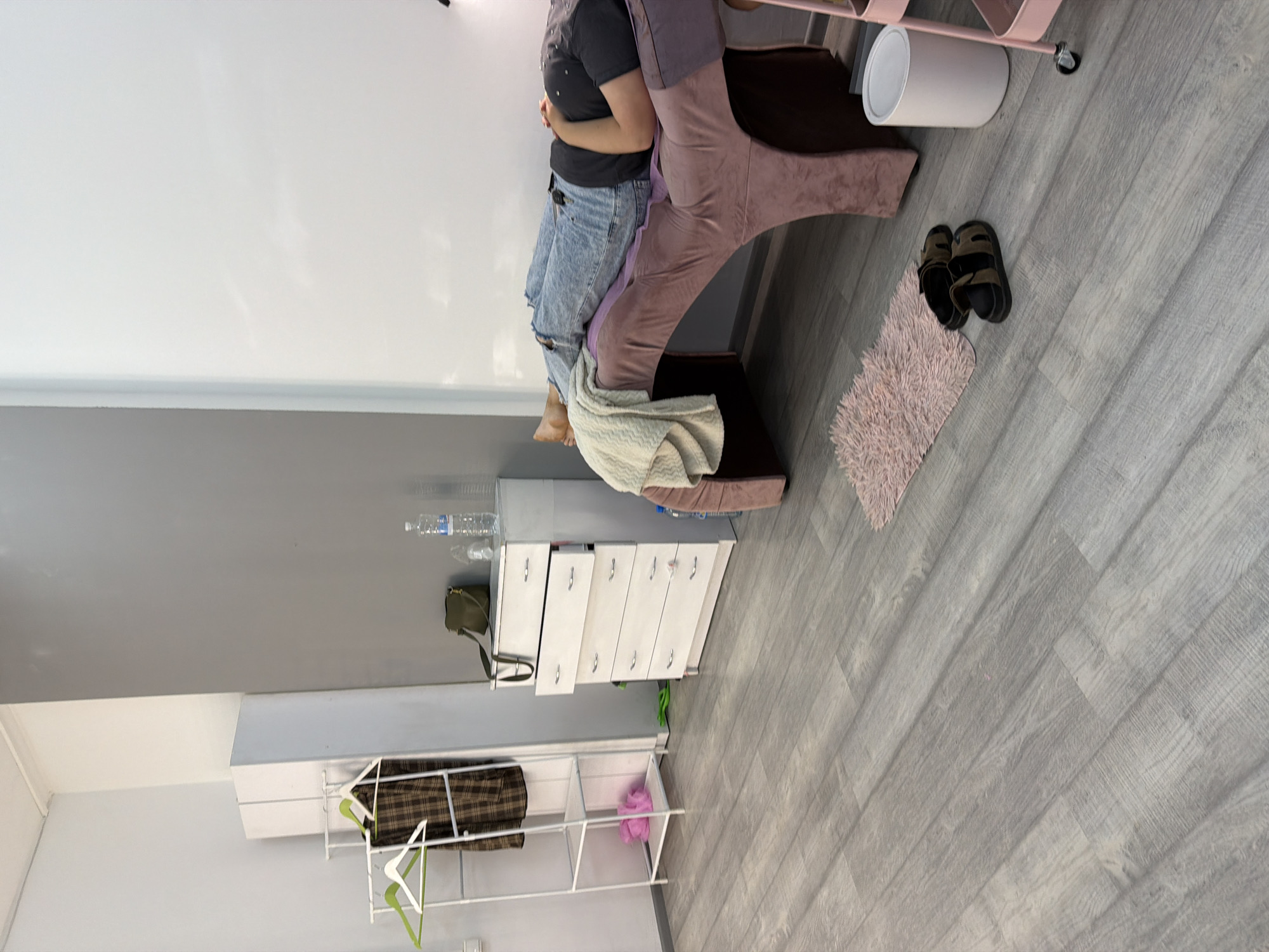 A person is lying on a pink cushioned table in a room with grey and white walls. The person is wearing blue jeans and a black t-shirt. A beige blanket is draped over their legs. To the left of the table, there is a white chest of drawers with a green purse on top. Next to the chest of drawers is a white clothing rack with a plaid garment hanging on it. On the floor, near the table, is a small pink rug and a pair of dark sandals. A white trash can is visible to the right of the rug, and part of a pink rolling cart is also in view. The floor is covered with grey laminate flooring. The overall scene suggests a private room, possibly for beauty treatments or relaxation, located in Kyiv, Ukraine.