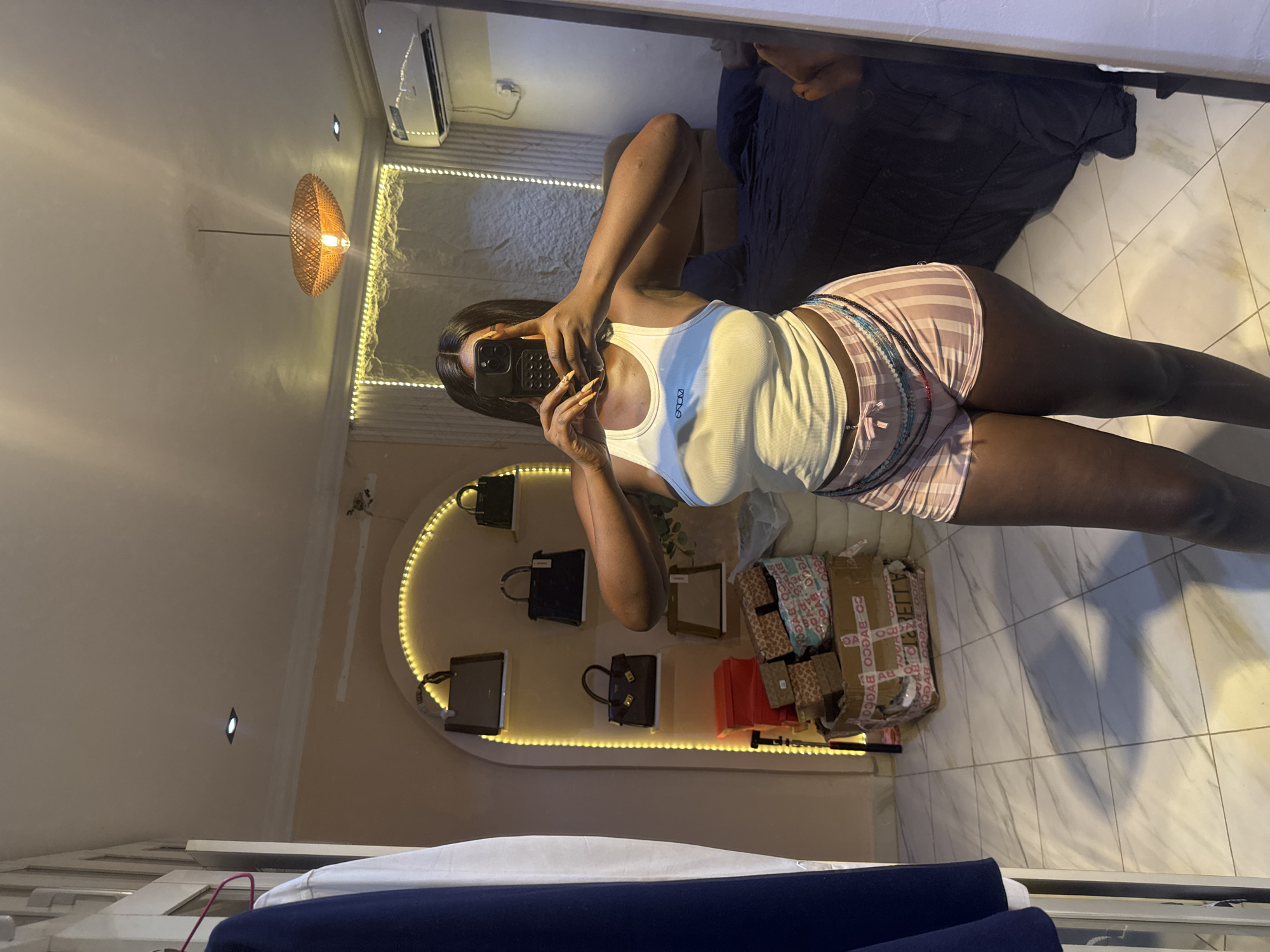 The image depicts an interior scene, likely a bedroom or dressing area, within a residence in Abuja, Nigeria. A young Black woman, taking a mirror selfie, is the central subject. She is wearing a white ribbed tank top and pink and white striped shorts. A beaded body chain is worn around her waist. The background reveals a tiled floor, a bed with dark blue bedding, and a wall with decorative lighting and shelves displaying several handbags. A large cardboard box with shipping tape is also visible in the room. The lighting suggests it is daytime, with artificial light sources also contributing to the illumination of the space.