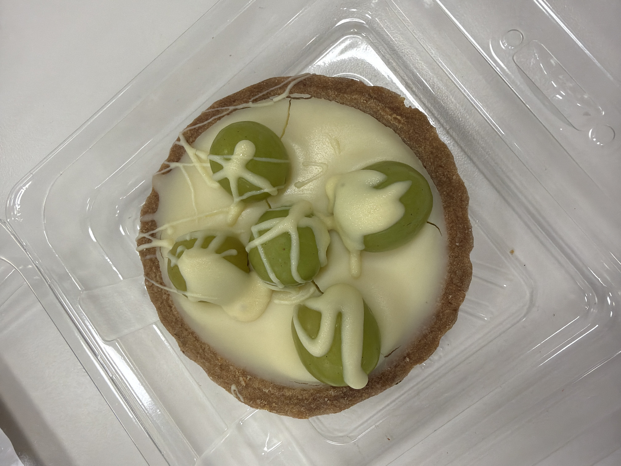 A top-down shot shows a small tart inside a clear plastic container. The tart has a crimped, light brown crust filled with a pale, creamy filling. Five green grapes are arranged on top of the filling, each drizzled with white chocolate or a similar topping. The container sits on a white surface. There is no contextual information to determine if this picture was taken in Puebla, Mexico. No people, actions, or interactions are visible.