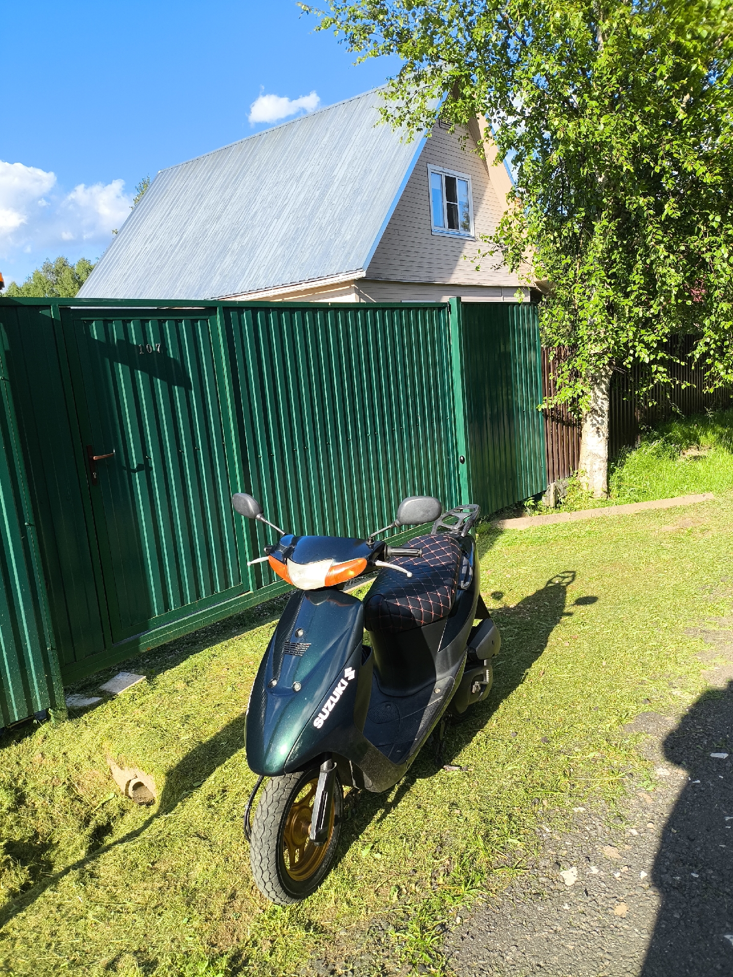 An outdoor, eye-level, medium shot captures a dark green Suzuki scooter parked on a grassy area next to a tall, dark green corrugated metal fence. The fence has a single metal handle and the number "107" is visible on it. Behind the fence, a light-colored, two-story house with a silver metal roof and a single window is visible. A large tree with green leaves is situated to the right of the house, partially obscuring it and casting shadows on the ground. The sky is blue with scattered white clouds, and the sun is shining. The ground is covered in green grass, with patches of cut grass near the scooter. A shadow of a person is visible on the right side of the image. The scene appears to be in a rural or suburban setting, potentially in the village of Trostyanka, Russia, given the context.