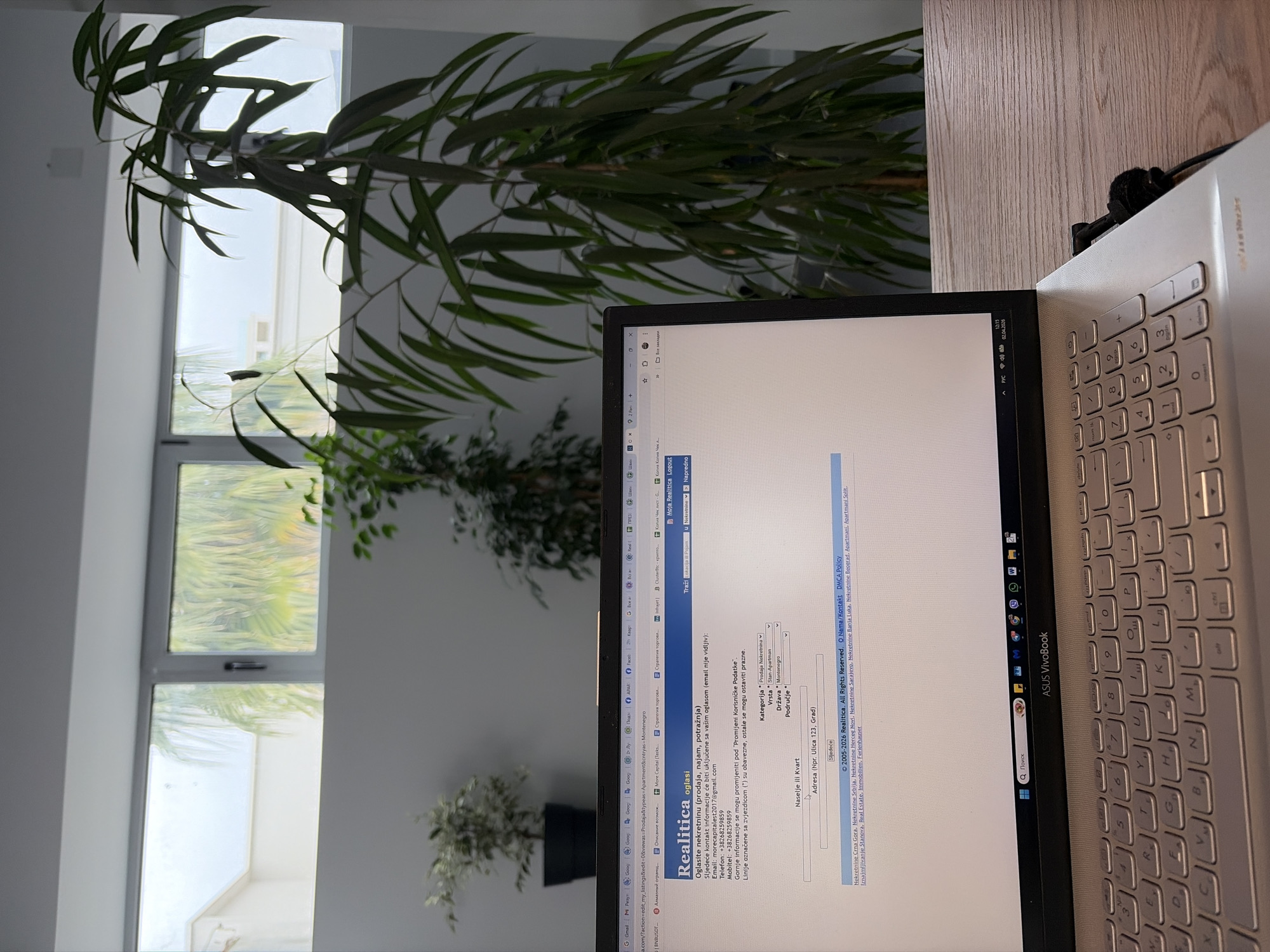 A photograph taken indoors, featuring a partially visible white laptop with an open web page displaying real estate listings in Montenegro, specifically mentioning apartments. The laptop screen is reflecting ambient light. The foreground is dominated by green foliage, suggesting an indoor plant. A window in the background reveals an exterior scene with greenery and what appears to be partial views of buildings, typical of coastal areas. Given the OCR data, the screen indicates the city of Budva, Montenegro is referenced. The scene suggests someone is working or browsing real estate options indoors, with a view of the city visible through the window.