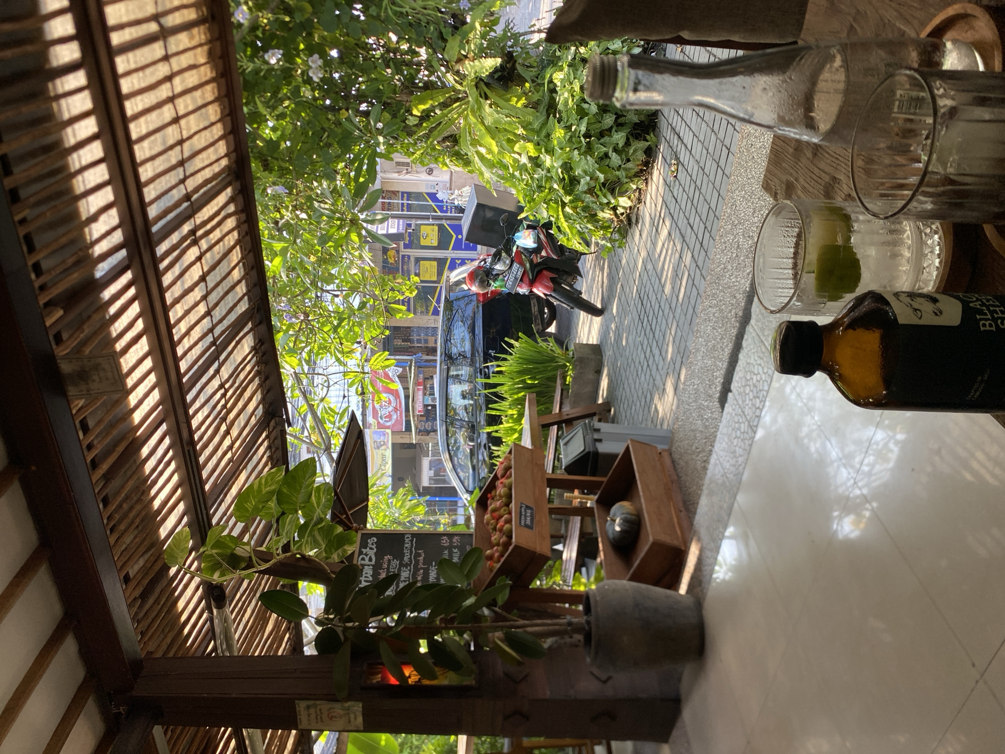 The image depicts an outdoor seating area of a cafe or restaurant in North Kuta, Indonesia. The foreground features a wooden table with two glasses of drinks with ice and lime, and a brown glass bottle with a black cap. To the left, a large potted plant stands next to a wooden pillar with a chalkboard menu. Behind the seating area, a paved walkway is visible, with a black car parked to the left and a red motorcycle with a black delivery box parked in the middle. Lush green foliage and trees surround the area, with a building facade visible in the background, displaying various signs and advertisements. The sunlight casts shadows through a slatted wooden roof overhead, suggesting it is daytime. The overall scene conveys a relaxed, tropical atmosphere.