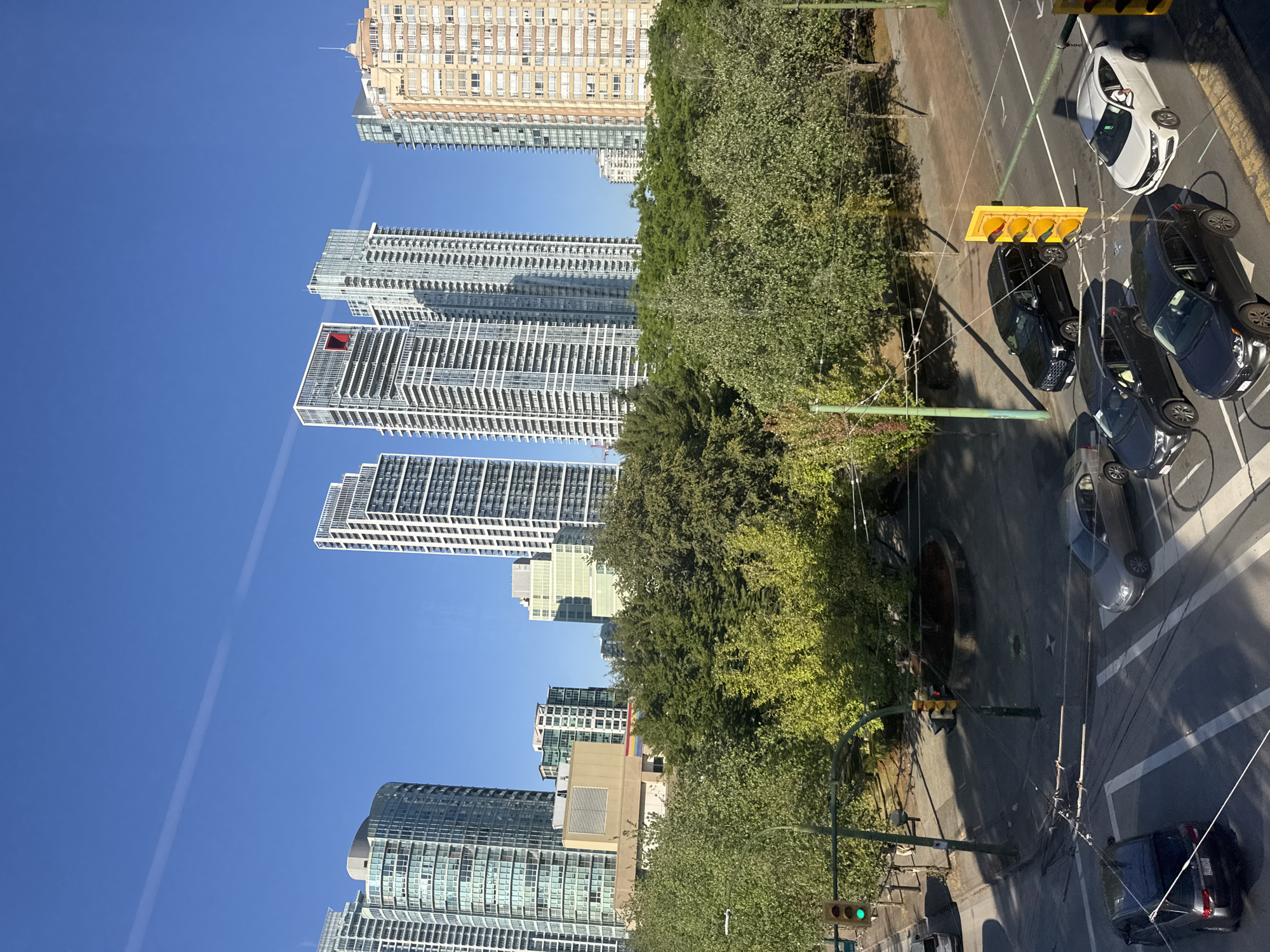 A high-angle, slightly tilted, full-shot photograph captures an urban street scene in Burnaby, Canada, under a clear blue sky with a faint contrail streaking across. The foreground features a section of a road with multiple lanes and pedestrian crosswalks. Several vehicles are visible: a dark SUV, two dark sedans, a silver sedan, and a white sedan. Traffic lights, one with a green signal illuminated, are positioned at an intersection. Power lines crisscross above the street.
Behind the road, a dense line of mature green trees forms a natural barrier. Beyond the trees, a collection of modern high-rise buildings dominates the mid-ground and background. These buildings vary in architectural style and height, with many featuring glass facades and white or beige exteriors. One prominent building has a distinctive red square design element near its top. Sunlight casts shadows across the scene, indicating it is daytime, likely late afternoon given the angle of the light. No people are clearly identifiable in the image. The overall impression is one of a bustling urban environment integrated with significant green spaces and modern architecture.