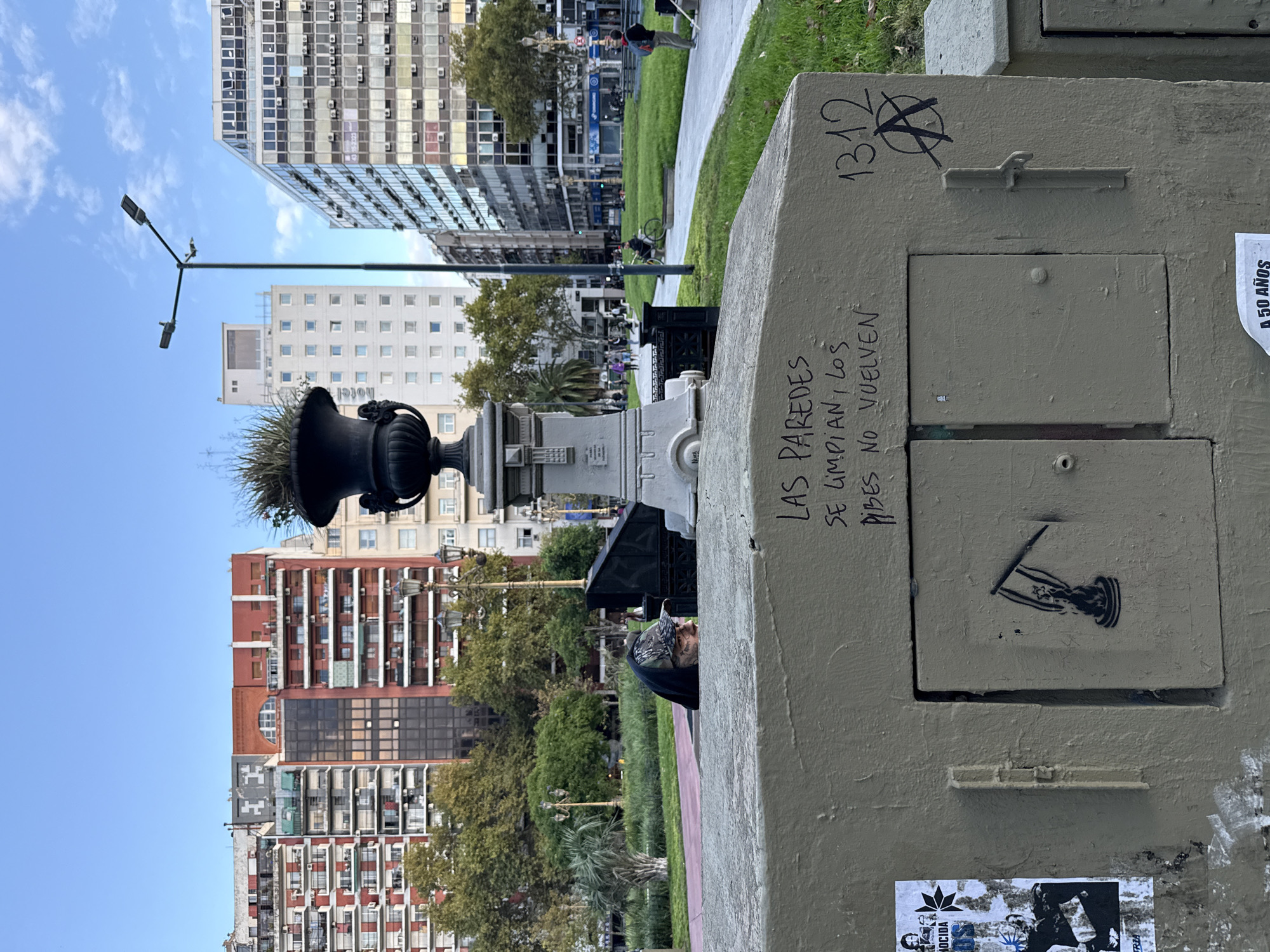 This photograph depicts a city park setting in Buenos Aires, Argentina. In the foreground is a weathered, grey structure with inscriptions and graffiti, including the number 1312 with an anarchy symbol. Graffiti inscriptions in Spanish read "LAS PAREDES SE LIMPIAN, LOS PIBES NO VUELVEN." There is a stenciled image of a figure with a flag and multiple stickers applied to its surface. Behind the structure, a person wearing a camouflage hat and a dark jacket is partially visible. A decorative urn with greenery sits atop a plinth behind the figure. In the background, there are several tall buildings of varying architectural styles, a street light, and a grassy lawn. People are walking along the pathways in the background. The sky is blue with scattered clouds.