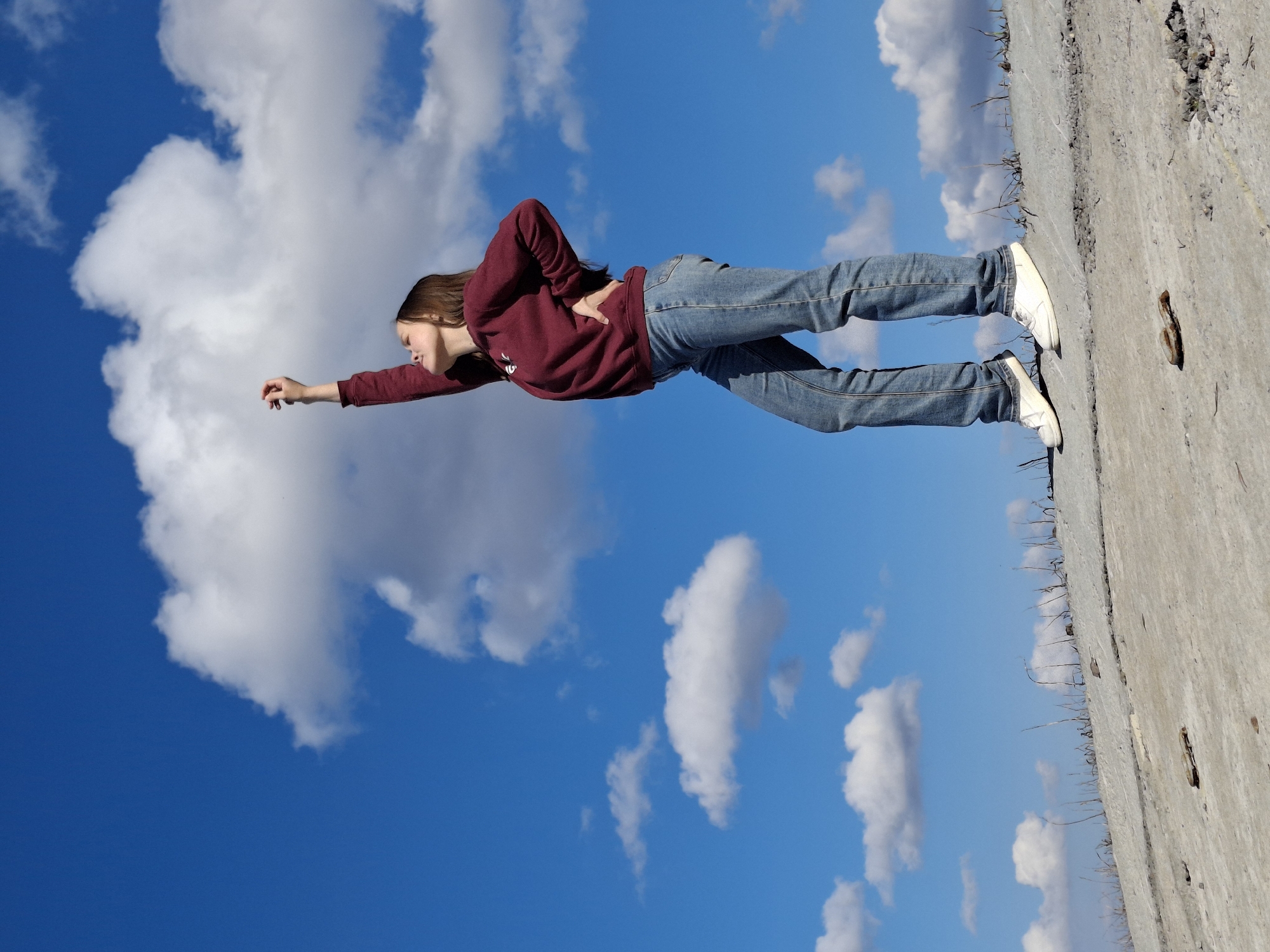 The image displays a young person appearing to fly horizontally across a vibrant blue sky dotted with fluffy white clouds. The subject, likely female, is dressed in a dark red long-sleeved top, blue jeans, and white sneakers. She holds a pose indicative of flight, with her body stretched out, one arm extended forward and the other hand on her lower back. Her head is tilted slightly, and her expression seems focused or serene.
The unique perspective of the photograph creates the illusion, as a concrete or paved surface runs vertically along the right side of the frame, serving as the ground the person is likely positioned on. This surface shows texture, small debris, and some dried vegetation. The lighting suggests it is daytime with clear, sunny weather. The overall impression is one of playful imagination and freedom. No text is visible in the image. The location context is Chaikovsky, Russia.