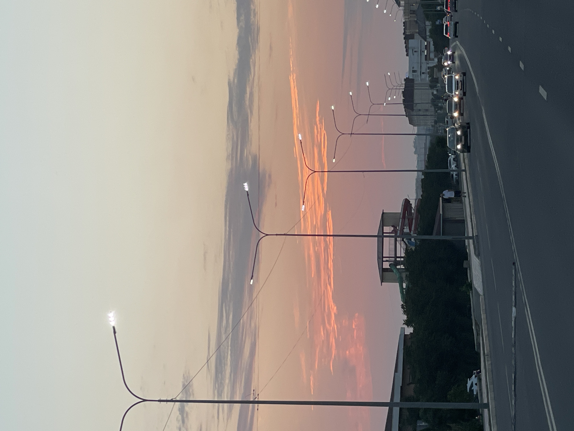 The image shows a wide asphalt road in Ömirzaq, Kazakhstan, with multiple lanes of traffic. Streetlights line the road, and several cars are driving in both directions. The sky is a gradient of pale blue, pink, and orange, suggesting it is either sunrise or sunset. On the left side of the road, there are buildings and a water park with slides. A few pedestrians are visible walking along the sidewalk. The overall scene depicts a typical urban environment at dusk or dawn.