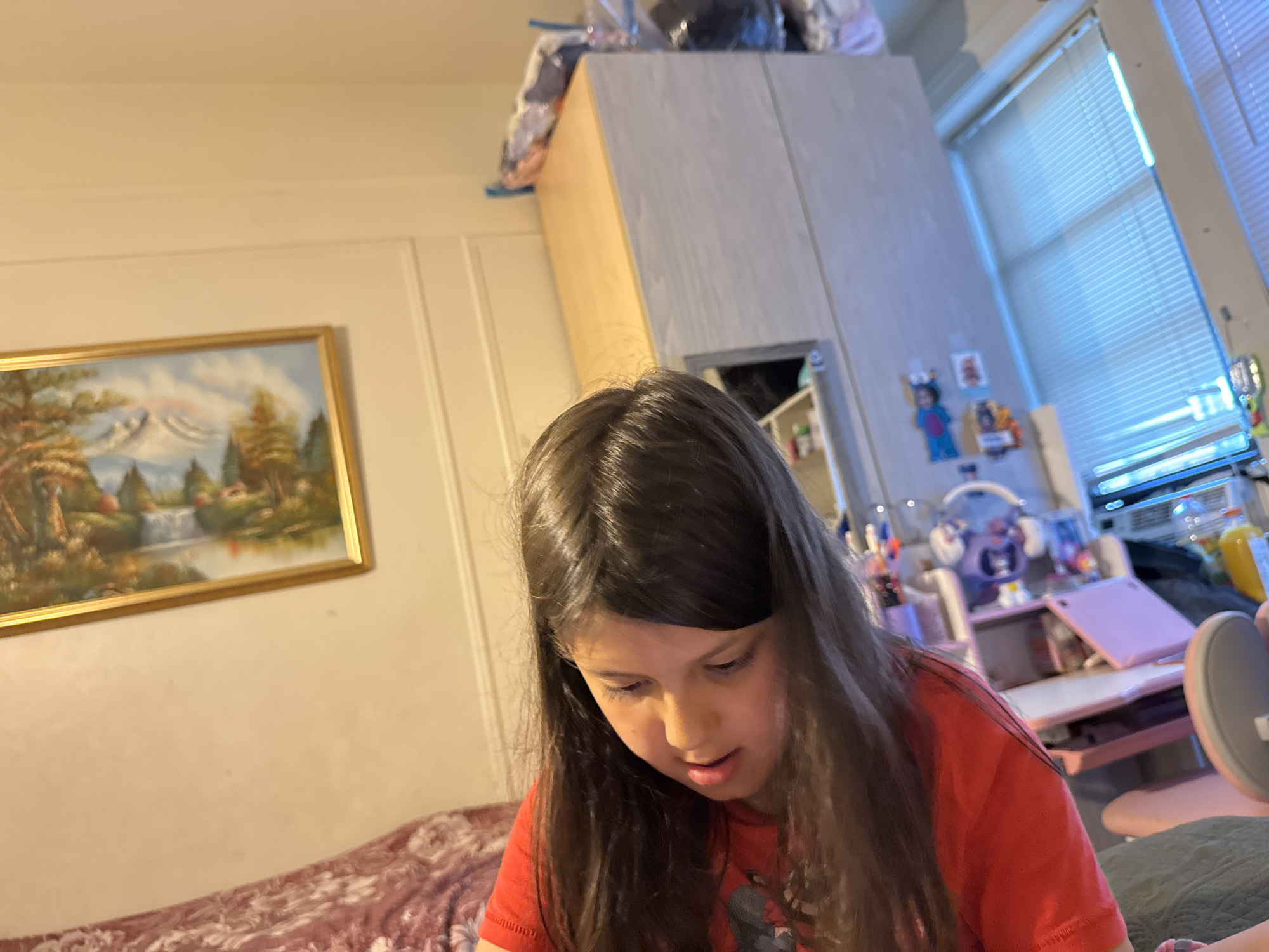 The image shows a young girl with long, dark hair, wearing a red t-shirt, looking down at something out of frame. She is in an indoor setting, with a painting on the wall to her left and a tall cabinet behind her. A window with blinds is visible on the right side of the frame. The scene appears to be a child's bedroom or play area. The image does not provide any specific information about the location in New York City, United States of America.