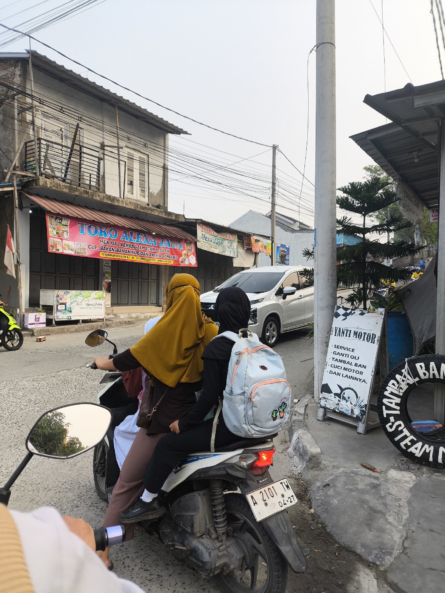 In Cilegon, Indonesia, on a street lined with modest buildings and shops, three individuals are observed on a motorcycle. The rider in the foreground, partially visible and wearing a white garment, holds the handlebars. Behind them, a person wearing a yellow hijab and a dark top is seated, with a brown bag slung over their shoulder. The third individual, wearing a black hijab and a backpack, sits behind them. A white SUV is parked further down the street, and a yellow motorcycle is seen to the left. A sign for "TOKO ALEXA JAYA" is prominently displayed on one of the buildings, advertising plastic goods, cake, and groceries. Another sign to the right indicates "YANTI MOTOR" services, including oil changes, tire repair, and car washing, with a large tire also present. The street is paved with asphalt and concrete, and overhead power lines crisscross the sky. The lighting suggests it is late afternoon or early evening.