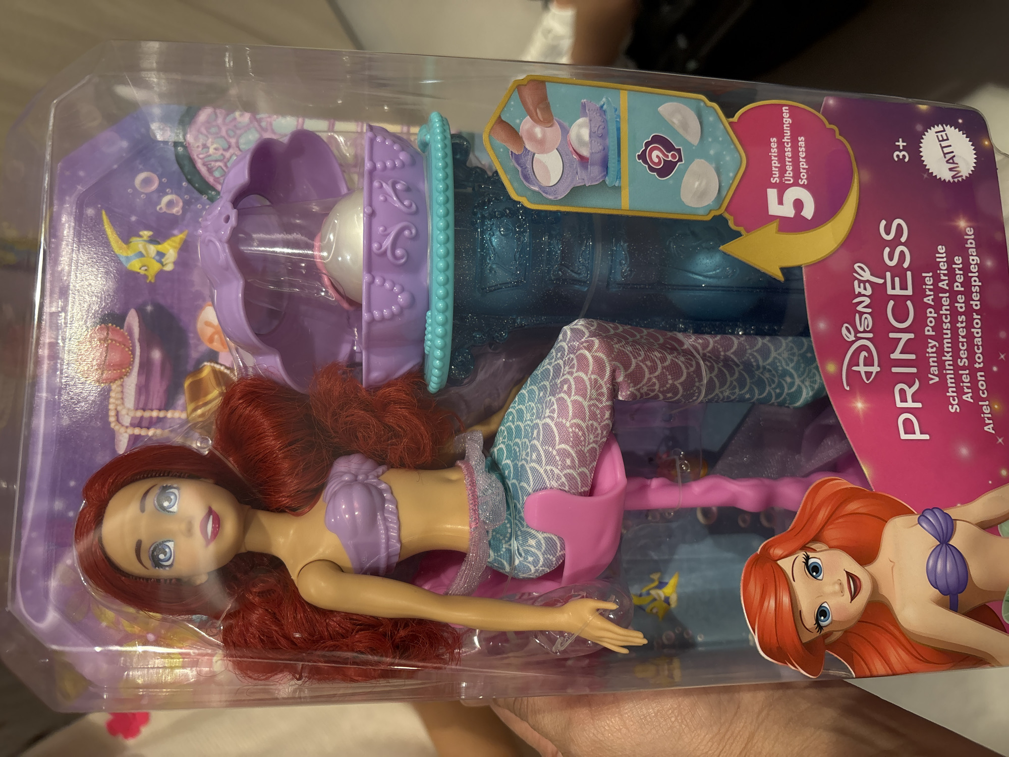 The image shows a Disney Princess Ariel doll in its packaging, a "Vanity Pop Ariel" toy. The doll is a mermaid with red hair, a purple seashell bra, and a blue and white scaled tail. She is seated on a pink stool. The packaging is clear plastic, revealing the doll and some accessories, including a purple vanity with a clam shell on top and a large pearl inside. There is also an illustration of Ariel on the front of the packaging, as well as text indicating that the toy includes "5 Surprises" and is suitable for ages 3+. The background of the packaging depicts an underwater scene with fish and bubbles. The image is a product photo of the toy and does not depict any specific location in Dubai, United Arab Emirates.