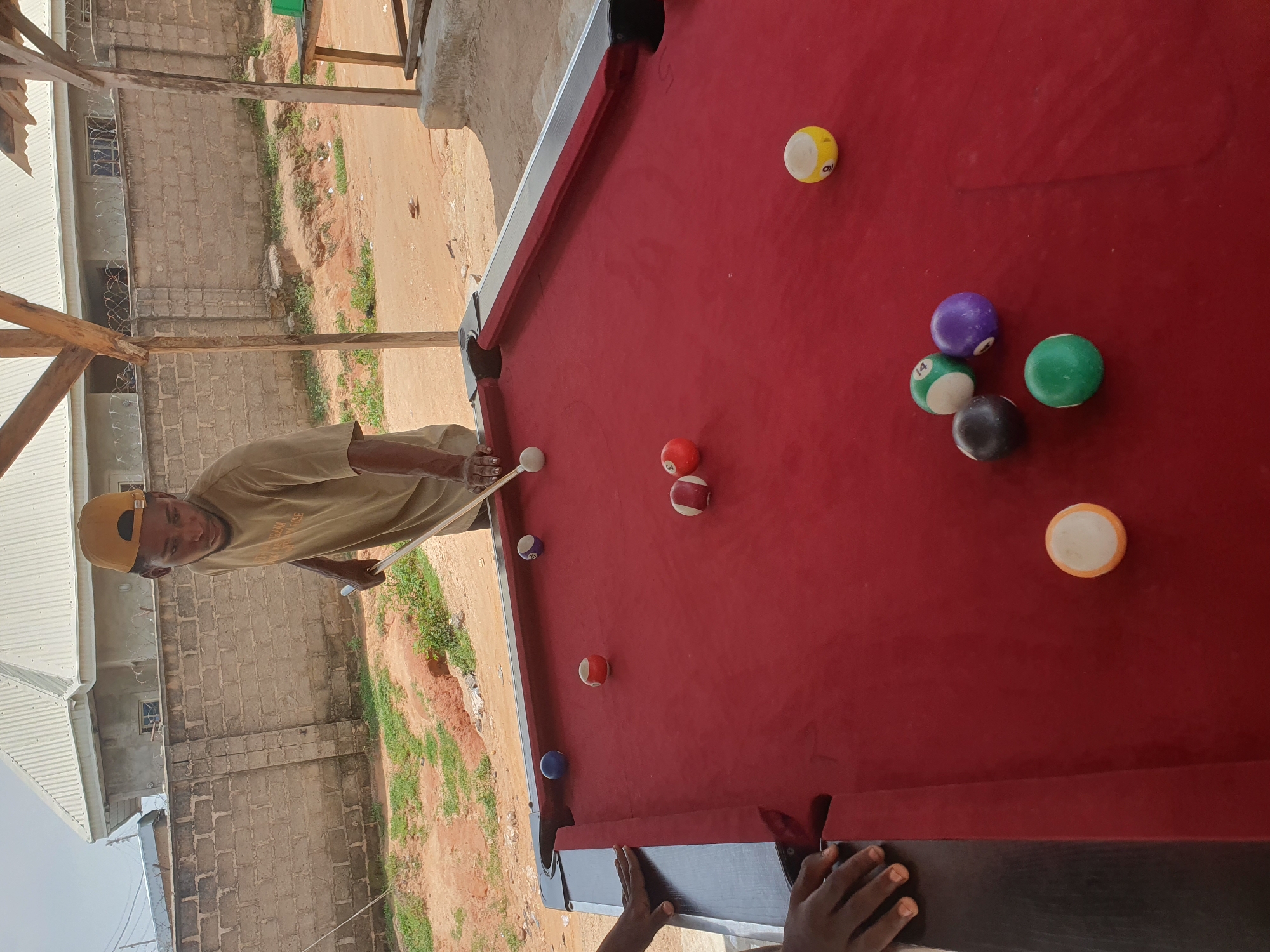A Black male wearing a yellow baseball cap and a tan t-shirt is playing pool on a red felt pool table. The pool table is outdoors, with a rough dirt and gravel ground surrounding it, and a grey brick wall and a light-colored building in the background. A wooden shelter with a corrugated metal roof is partially visible. There are several billiard balls scattered across the table, including a white cue ball, a blue ball, a red and white striped ball, a solid red ball, a solid purple ball, a solid black ball, a green ball with a white stripe, a green ball with a white stripe and the number 14 on it, and a yellow and white ball with the number 9 on it. The man is holding a pool cue and appears to be in the process of taking a shot. The scene suggests a casual game of pool in an informal outdoor setting in Lokoja, Nigeria.