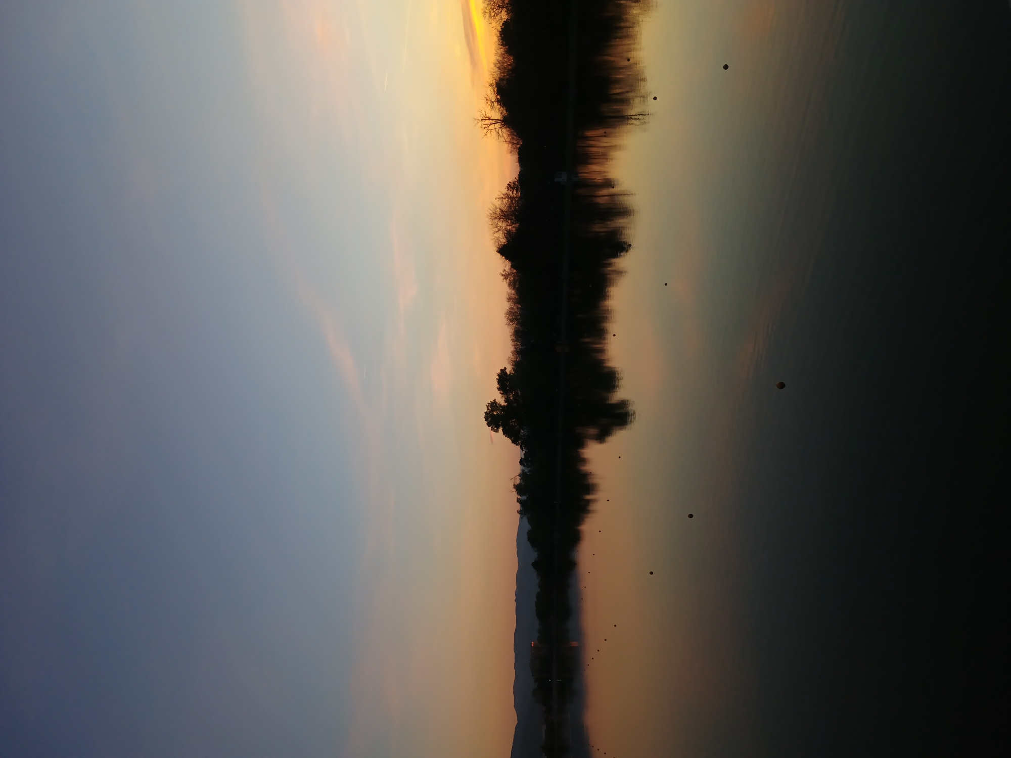 The image, taken in Plovdiv, Bulgaria, captures a serene lake or calm body of water during either sunset or sunrise. The sky displays a beautiful gradient, transitioning from a dark indigo-blue on the left to warm, vibrant shades of orange and yellow towards the right. Faint, wispy clouds and a subtle contrail can be observed in the lighter parts of the sky.
A distinct line of silhouetted trees and distant mountains stretches across the horizon, sharply contrasting with the colorful sky. These natural elements are perfectly mirrored in the still water below, creating a powerful reflection that duplicates the sky's color palette and the dark landforms.
Several small, dark, circular objects, appearing to be buoys, are visible floating on the water's surface, particularly in the middle ground and extending towards the darker foreground on the right. The calm conditions are evidenced by the clear and undisturbed reflections. The overall scene conveys a sense of tranquility and natural beauty during the golden hour. No visible text is present.