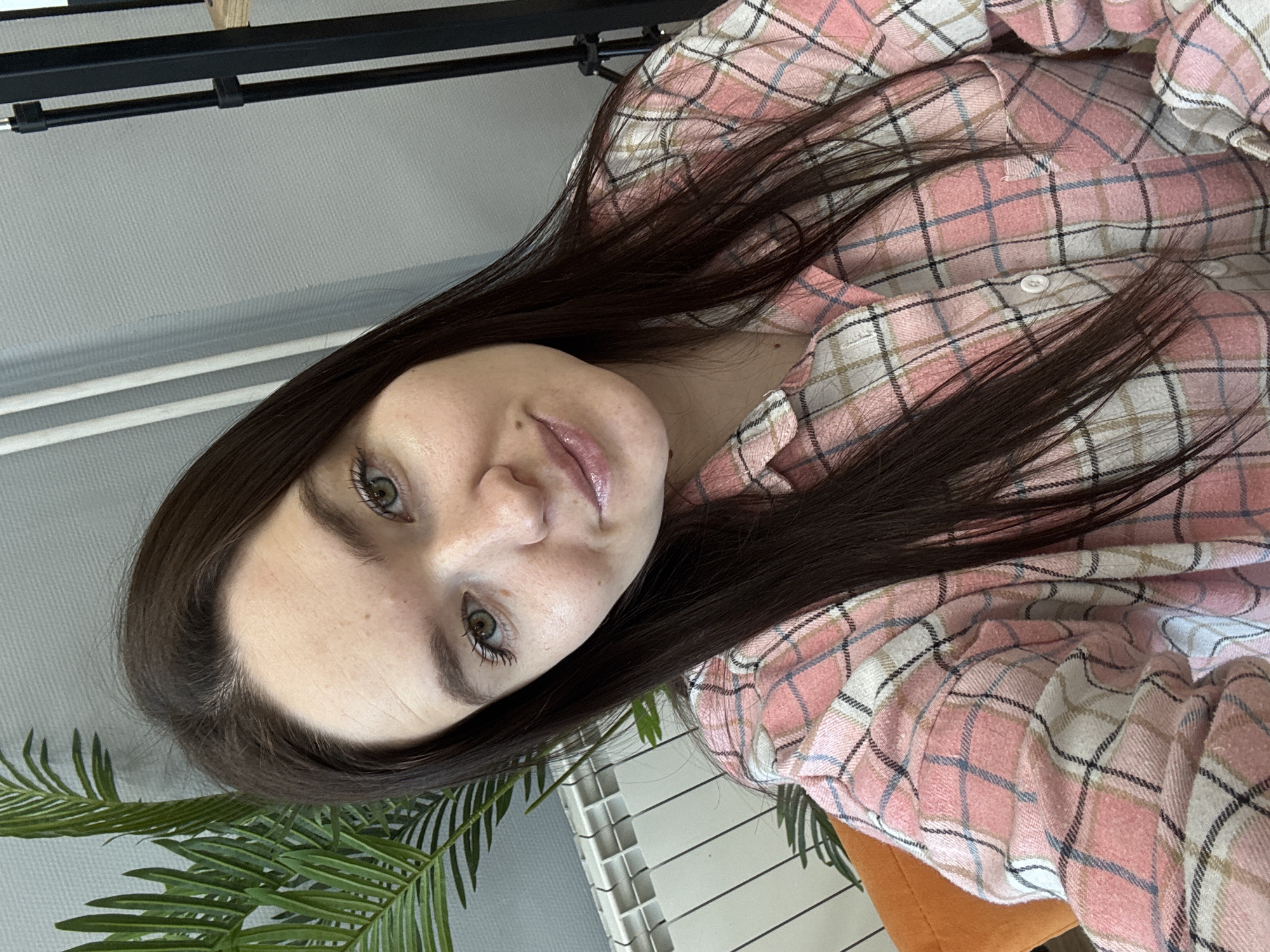 A young woman with long, dark brown hair and fair skin is shown in a close-up, angled headshot. She has light-colored eyes and is wearing a pink and white plaid flannel shirt. Part of a green, leafy plant is visible in the lower left corner of the frame. The background includes a light gray wall, a white radiator, and a black metal frame. The location is Chaikovsky, Russia.