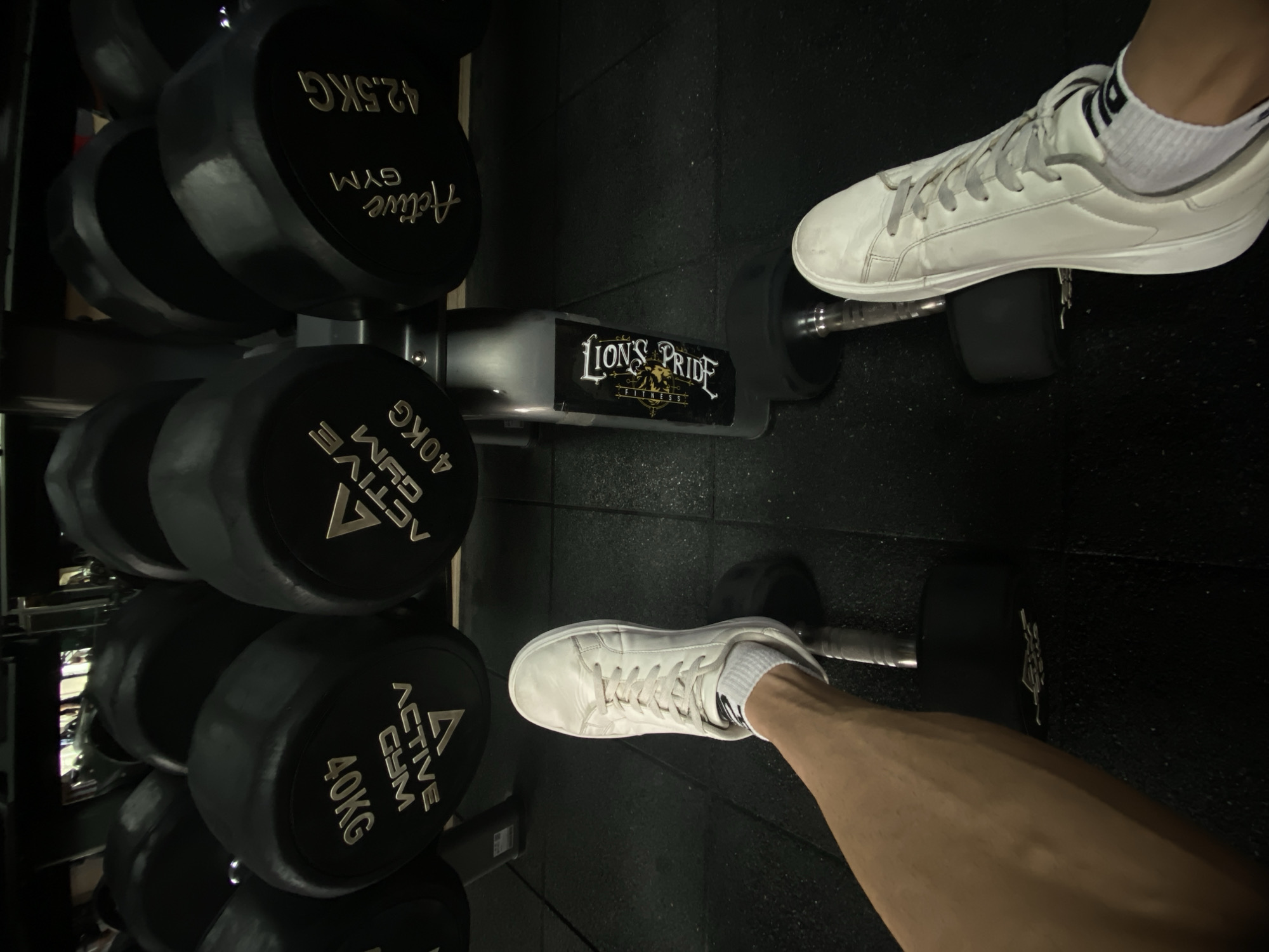 The image shows a gym environment with a rack of dumbbells ranging from 40kg to 42.5kg labeled "Active GYM". In the foreground, there are two dumbbells on the floor, each partially obscured by a person's foot. The person on the right is wearing a white shoe and white sock, while the person on the left is also wearing a white shoe with a white sock and a portion of their leg is visible. A sticker on one of the dumbbell holders reads "LION'S PRIDE FITNESS". Based on the branding of the dumbbells and the fitness studio name, it is likely that this image was taken at a fitness center within Pleven, Bulgaria.