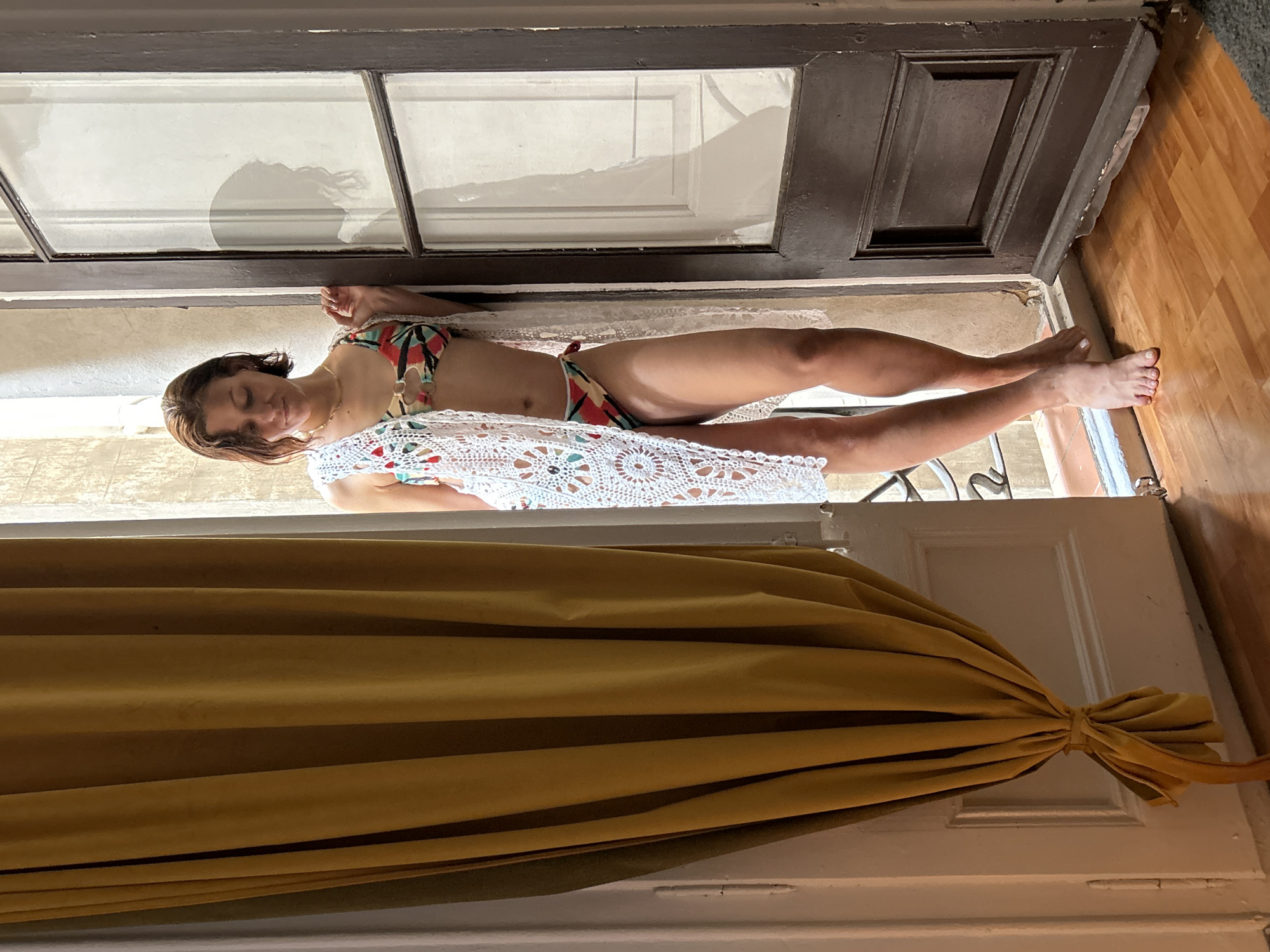 A woman in a swimsuit lies diagonally across a doorway or entrance, partially covered by a white crocheted blanket. The doorway is framed by a dark-colored frame and leads to a room with wooden flooring. A large, draped mustard-colored curtain hangs below the doorway. The light suggests an interior setting. Barcelona, Spain, is not verifiable from the image.
