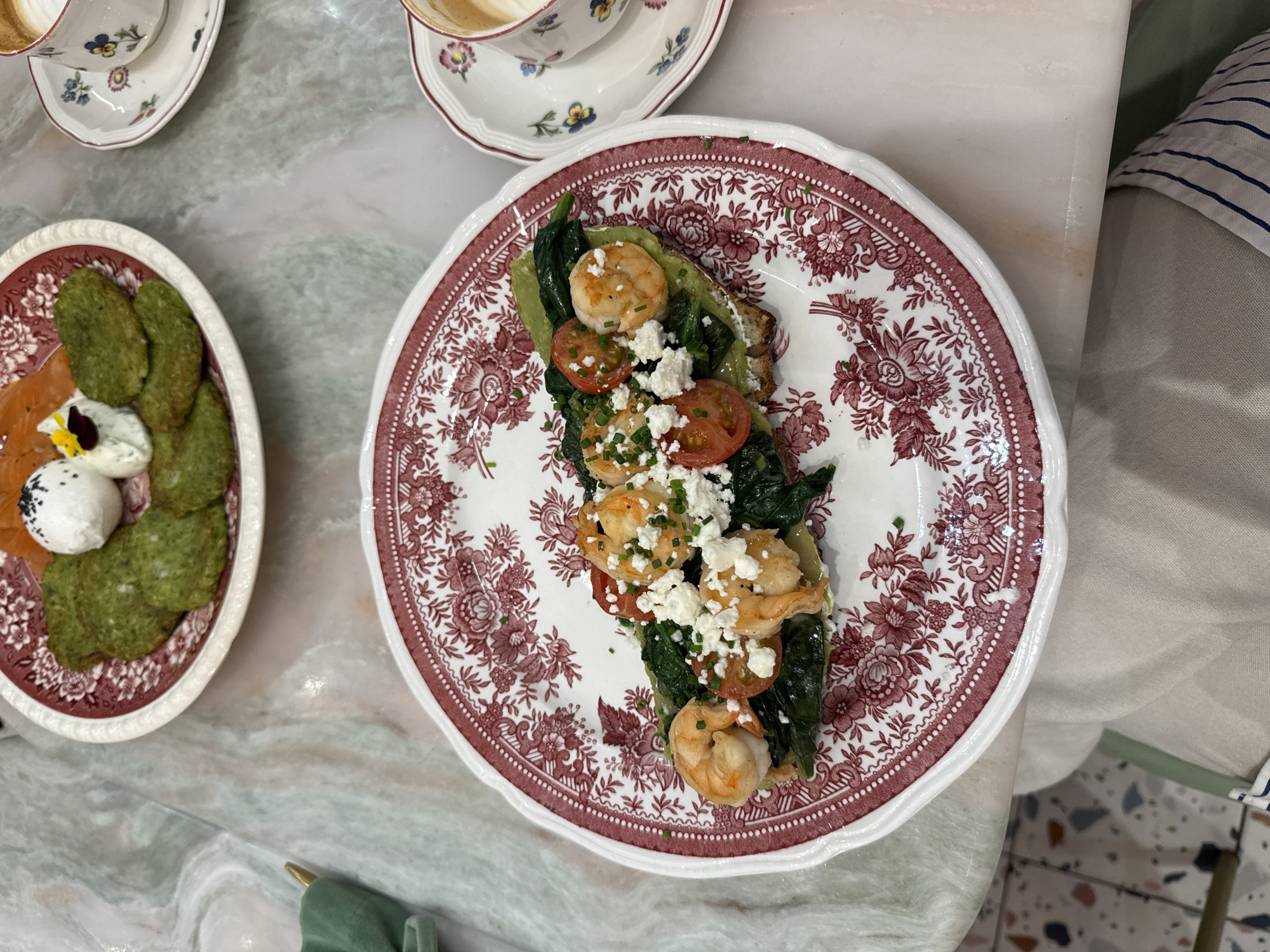 A close-up, top-down shot captures a brunch spread on a marble-patterned table. The main focus is a white plate with a red floral design, featuring an open-faced sandwich. The sandwich is topped with mashed avocado, wilted spinach, halved cherry tomatoes, crumbled feta cheese, and several grilled shrimp. Chopped chives garnish the top. To the right, a teacup and saucer with a similar floral pattern hold a latte. In the background, another plate displays green patties, a small mound of white cheese with black seeds, and slices of smoked salmon. The overall scene suggests a breakfast or brunch setting, possibly in a cafe in Kyiv, Ukraine.
