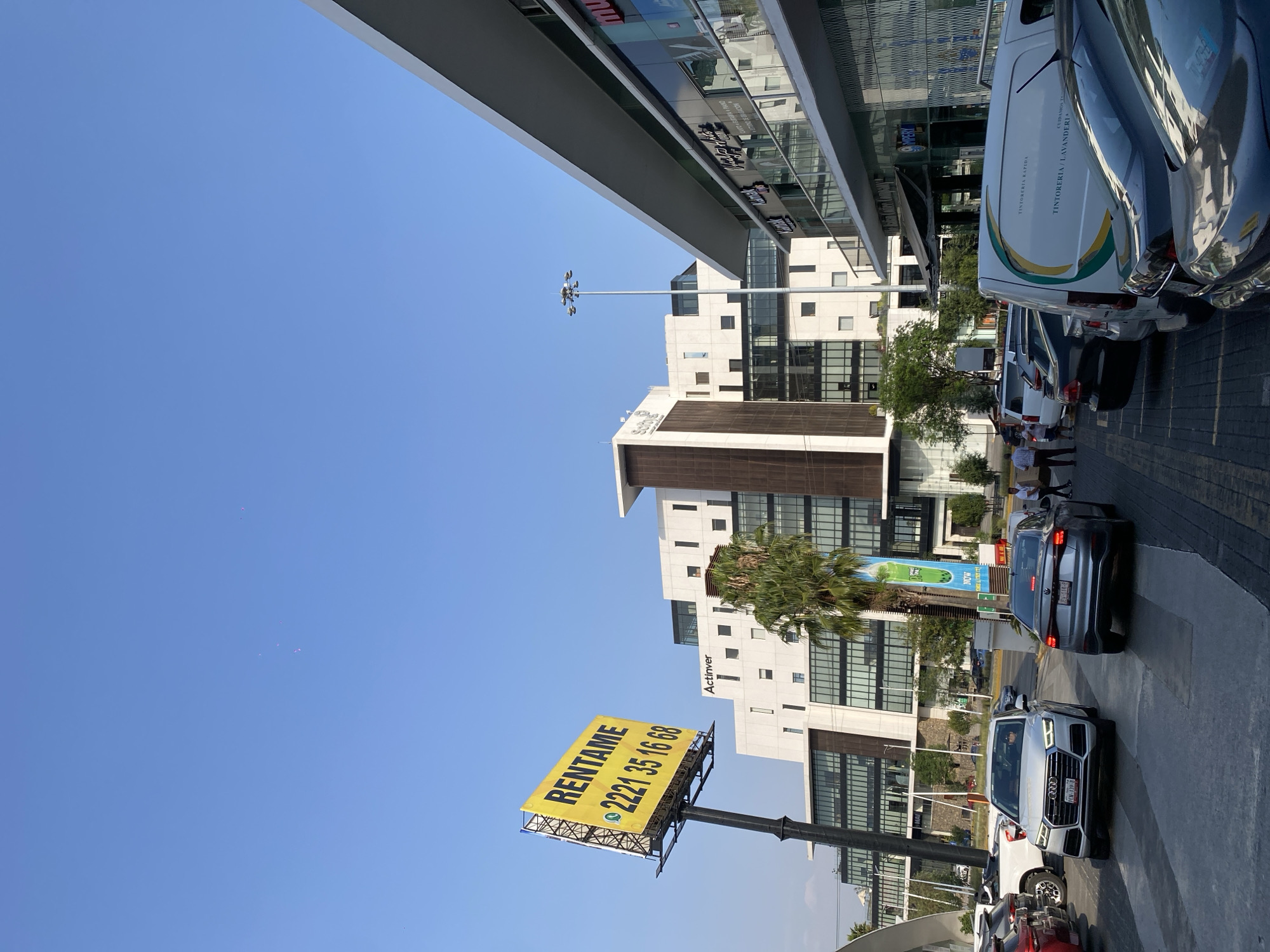 The image captures an urban scene with several notable elements. The sky above is clear and blue, suggesting a sunny day. A tall commercial building or shopping center with a modern design is on the left, featuring glass and metal accents.
A large billboard stands prominently on the right, displaying the word "RENTAME" (rent me) and a phone number. This suggests it is an advertisement for rentals. In the background are white buildings with black window frames and a palm tree.
The foreground contains a line of parked cars. A white Audi SUV is the most visible car.
The environment appears to be a busy commercial area with a mix of shopping, office, and residential buildings. The location is unknown.