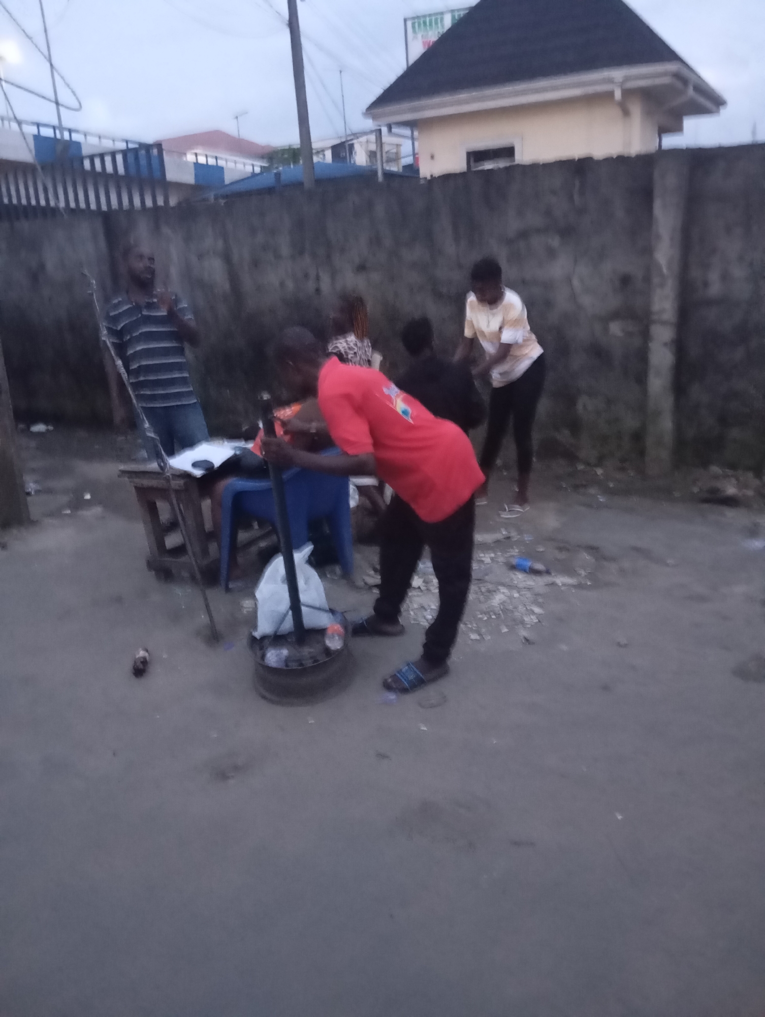 Four individuals, two male and two female, are gathered around a makeshift stall. The stall consists of a wooden table with papers on it, a blue plastic chair, and a large circular metal object on the ground, likely a cooking pot or brazier, with a white plastic bag on top and a metal rod inserted into it.
One male, wearing a striped t-shirt and dark trousers, stands behind the table. Another male, in a red t-shirt and dark trousers, is bent over the metal object on the ground, appearing to adjust or tend to something within it. A female wearing a light-colored t-shirt with horizontal stripes and dark trousers stands to the right of the male in red, looking towards him. A fourth individual, a female with braided hair, is partially visible behind the male in red, wearing a patterned top.
The environment appears to be a street or roadside in Port Harcourt, Nigeria. The ground is unpaved and dusty, with scattered debris. A concrete wall runs along the background, with a fence above it. A building with a dark-tiled roof is visible behind the wall. The lighting suggests it is either early morning or late evening. Several bottles and other small discarded items are visible on the ground. The overall scene depicts a candid moment of activity at a street-side setup.