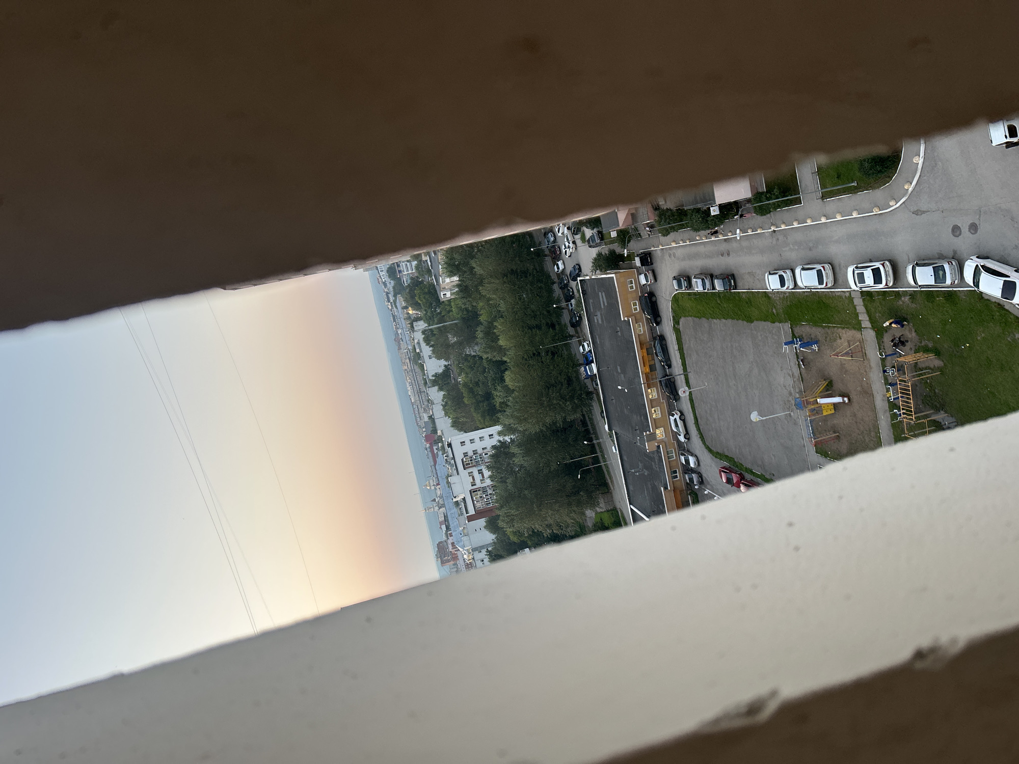 The image shows an elevated view of a residential area in the city of Perm, Russia, likely during sunset due to the warm hues in the sky. Two large, textured architectural elements frame the scene, obscuring parts of the view on the left and right.
In the foreground, a children's playground with yellow and red structures is visible, situated on a sand-covered area. To the right of the playground, a paved road curves through the scene, lined with parked cars of various makes and models, predominantly white and silver sedans. Further along the road, a few pedestrians can be seen walking.
Beyond the playground and road, several multi-story apartment buildings are scattered amidst a dense canopy of green trees. Some of these buildings have visible windows and balconies. In the distance, a body of water, possibly a river, stretches across the horizon, with more buildings and structures visible on its far bank, suggesting a larger urban landscape. Power lines are also present in the sky. The overall impression is a typical urban scene with residential blocks, green spaces, and infrastructure.