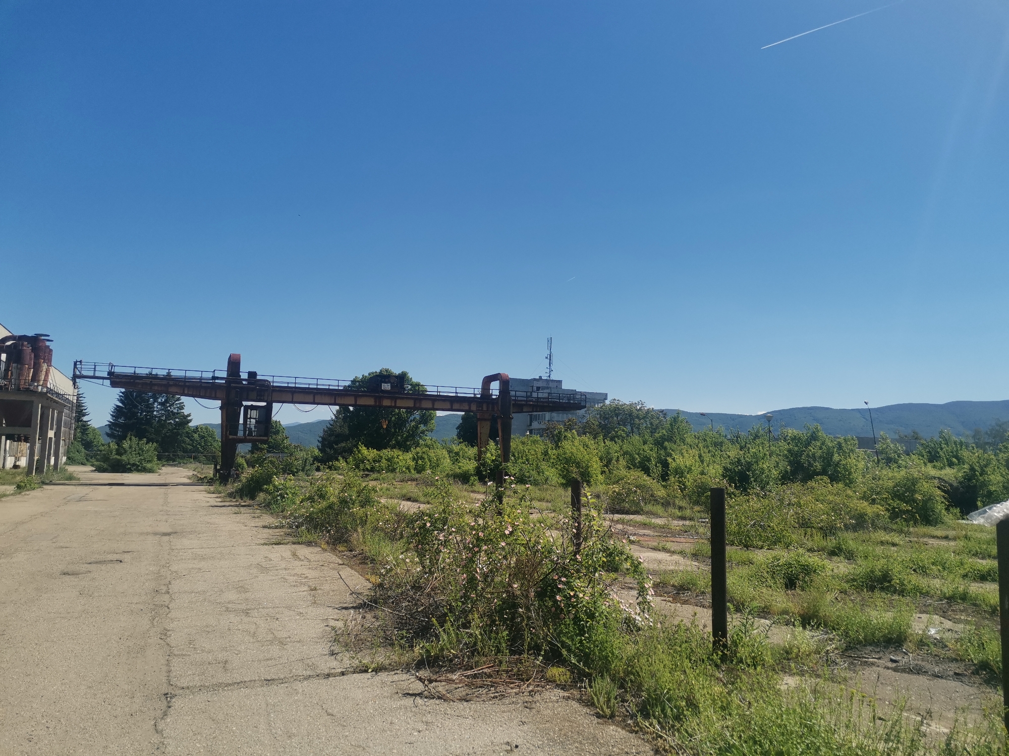 The image depicts an abandoned industrial site in Zhivkovo, Bulgaria. A cracked concrete road extends into the scene, flanked by overgrown vegetation. An overhead crane structure spans the road, showing signs of rust and disuse. In the background, a building with an antenna is visible, along with distant mountains. The sky is clear and blue, with a contrail from an aircraft.
