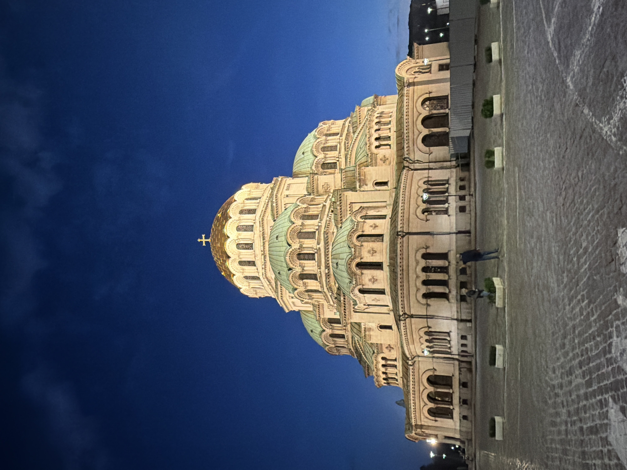 The image depicts the St. Alexander Nevsky Cathedral in Sofia, Bulgaria, at dusk or night. The cathedral occupies the majority of the midground, illuminated against a deep blue sky. Its architecture is characterized by a large central golden dome topped with a cross, surrounded by numerous smaller domes with a green patina finish. The building's facade is light-colored, featuring multiple tiers of arched windows and decorative elements.
In the foreground, a cobblestone plaza extends towards the cathedral. Several rectangular white planters containing small green bushes are evenly spaced across the plaza. Two individuals are standing on the plaza in the mid-left portion of the frame; one person is positioned closer to the viewer and the other slightly further back and to the right, both facing towards the cathedral. Their attire appears dark, and their specific actions are indistinct.
The sky above the cathedral is a dark, clear blue, with subtle cloud formations visible in the upper left quadrant. Artificial lighting from streetlights or adjacent structures is discernible in the far background on both the left and right sides. On the far right, behind the planters, a section of what appears to be temporary fencing or construction barriers is visible. The overall scene is well-lit, highlighting the cathedral's features against the dark sky.