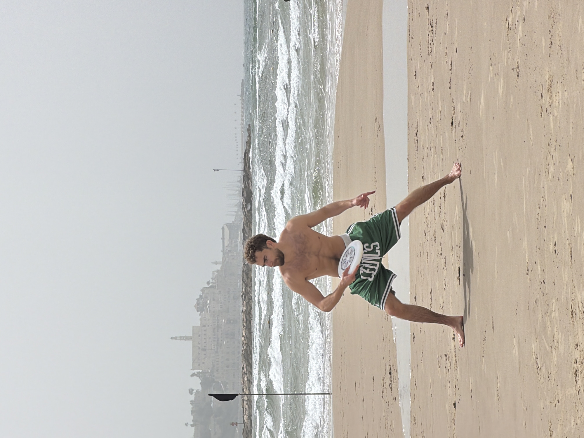 A man is seen on a sandy beach next to the Mediterranean Sea in Tel Aviv, Israel. The man is fair-skinned, shirtless, and wearing green shorts with "CELTICS" written on them. He is holding a white frisbee and appears to be about to throw it. The beach transitions into foamy waves and then the sea. In the distance is a skyline of Tel Aviv, obscured slightly by haze. In the middle ground, a tall pole stands near the water's edge with a black flag attached.