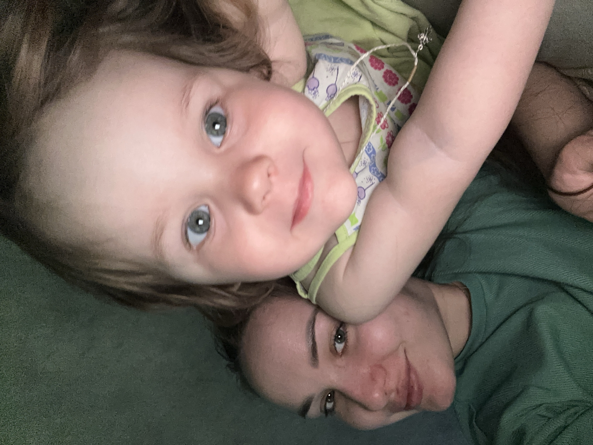 A close-up, indoor shot depicts a young child lying on top of an adult. Both are facing the camera. The child is on top, slightly angled, wearing a light-colored sleeveless top with a floral pattern. The adult is underneath, wearing a dark green shirt. The child's arm is draped across the adult's shoulder. The background is a solid green color, possibly a couch or bedding. The specific location in Chaikovsky, Russia, cannot be determined from the image.