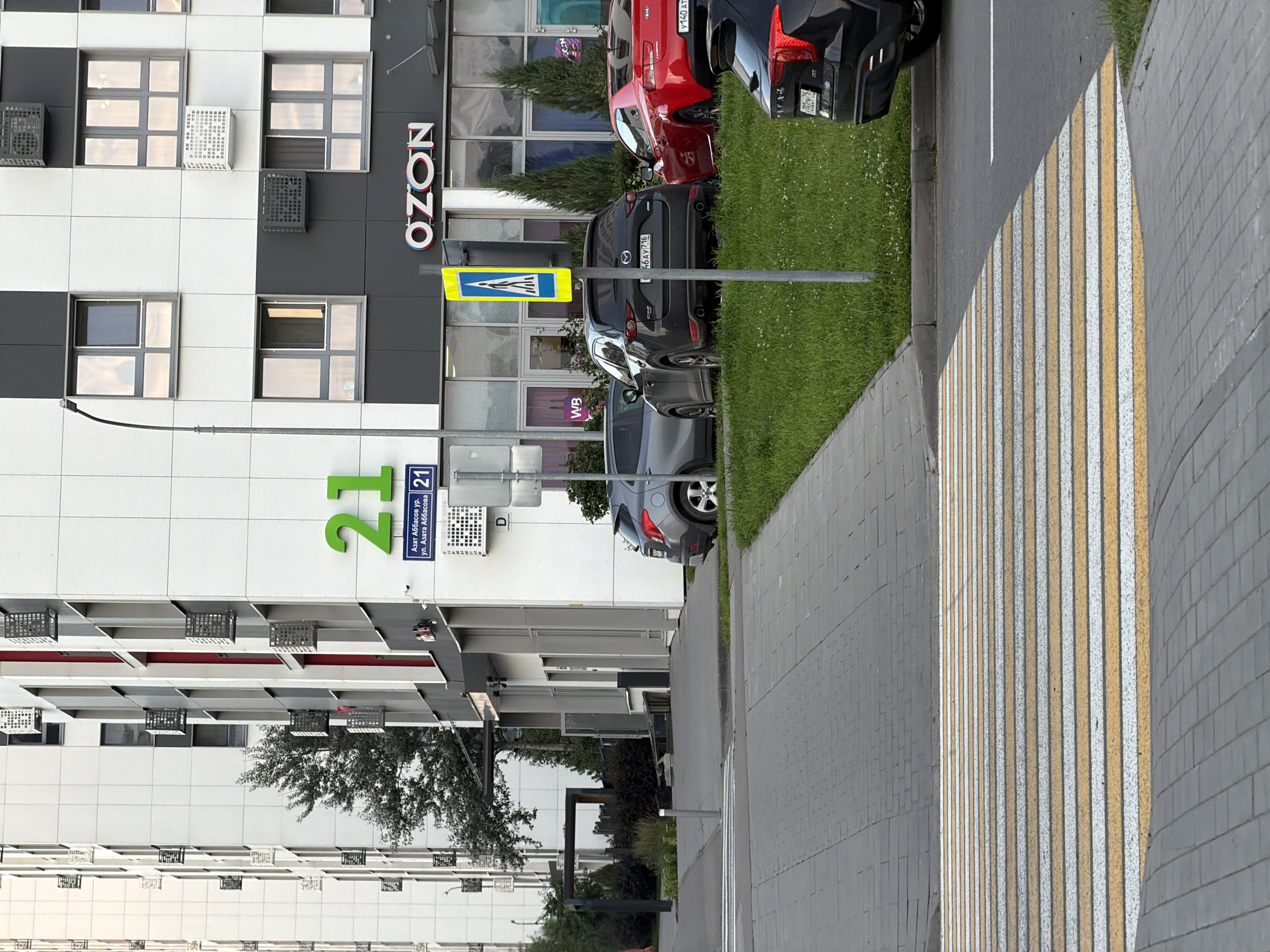 The image shows a street scene in front of a modern apartment building. The building has a white facade with gray accents and balconies. The building's address, "21," is displayed prominently in large green numerals. Below the number "21," a street sign reads "Азат Аббасов ур. ул. Азата Аббасова." A pedestrian crossing is visible in the foreground, with yellow and white stripes painted on the asphalt. Several cars are parked along the street, including a dark gray Mazda CX-5, a red Kia, and a black SUV. A bright yellow pedestrian crossing sign is posted on a metal pole. The environment appears to be an urban setting with a sidewalk, grass, and trees.