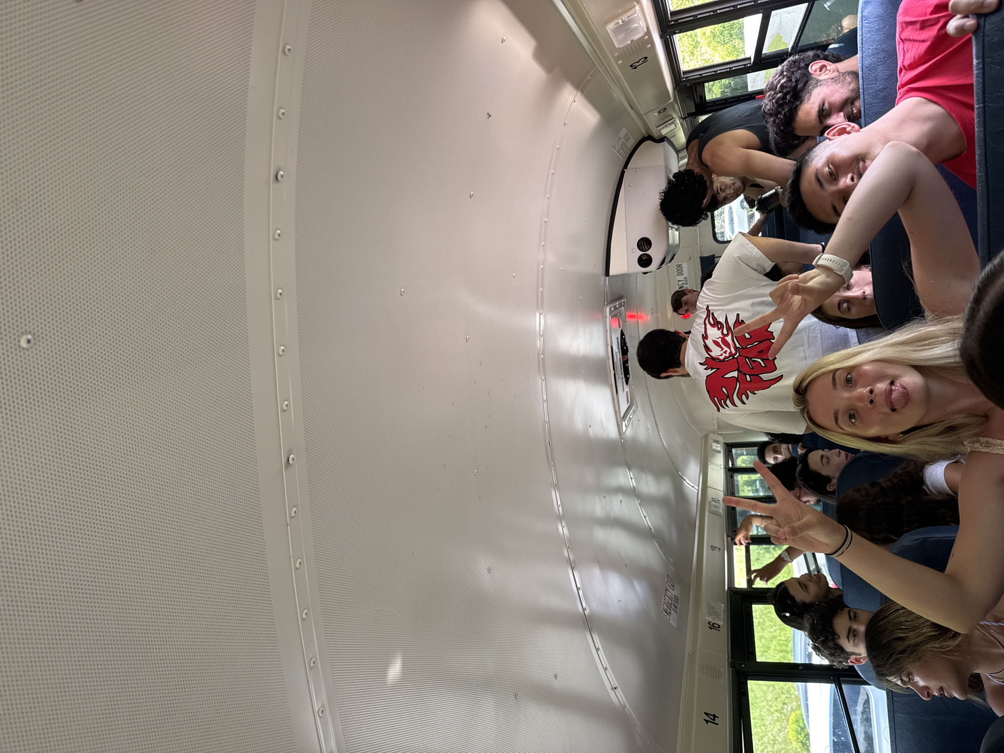The image depicts the interior of a school bus filled with young people, likely on a field trip or excursion. The bus is in motion, as indicated by the blurry movement of the exterior foliage visible through the windows. The people are predominantly young adults, dressed casually. Some are looking at the camera, while others are engaged in conversation or looking out the windows. The bus ceiling is white and textured, with visible seams and lights. There are overhead storage compartments and ventilation systems. Numbers indicating seat rows (13, 14, 16, 18, 20) are visible on the side of the bus. One person in the foreground is making a peace sign with their fingers and sticking out their tongue. Another person, wearing a white t-shirt with a red graphic, is also making a peace sign. The overall atmosphere appears to be lively and informal. The specific location in Washington Township, United States of America is not identifiable from the image.