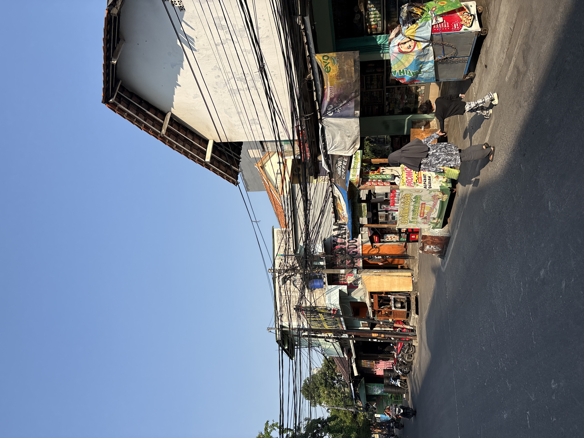 This image depicts a street scene in North Jakarta, Indonesia, likely on a bright, sunny day with a clear blue sky. The street is lined with a mix of small businesses and food stalls, some with awnings and signage in Indonesian. The buildings appear to be a mixture of residential and commercial, with several stories. Overhead, a dense network of black electrical wires creates a tangled web across the scene, a common sight in urban Indonesian environments.
In the foreground, two people, a woman wearing a hijab and a child, walk away from the camera down the street. The woman is dressed in a patterned grey and black outfit, and the child, in a black shirt and patterned pants, holds her hand. They are casting long shadows on the asphalt road. To their right, a colorful food cart is parked, covered with various tarps and signage, suggesting it is used for selling goods.
Further down the street, more food stalls and shops are visible, with motorcycles parked in front of some. The architecture is modest, with visible roofs and window frames. The overall impression is of a busy, functional urban environment with everyday street life unfolding.