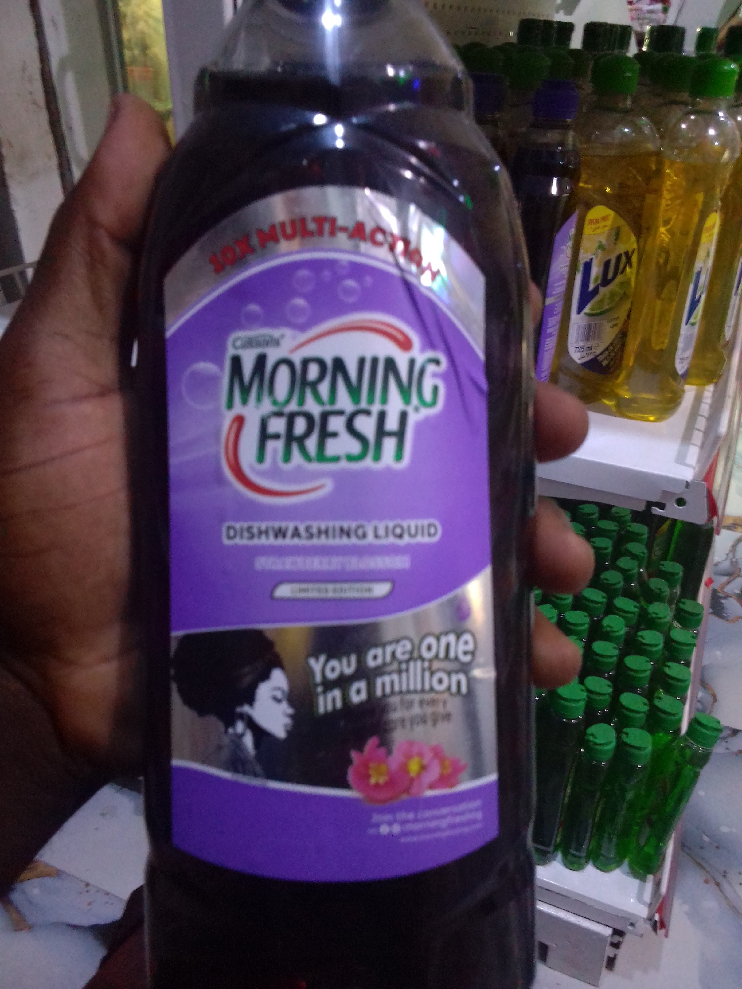 A person's hand is holding a large bottle of "Morning Fresh Dishwashing Liquid." The bottle has a purple label with a silver band at the top. The label features the "Morning Fresh" logo in green and white, along with an illustration of a woman's profile and pink flowers. The text on the label includes "10X MULTI-ACTION" and "DISHWASHING LIQUID". In the background, on shelves, are rows of smaller green-capped bottles, some of which appear to be Lux dish soap. The scene appears to be in a store or market setting. The lighting is artificial and somewhat dim.