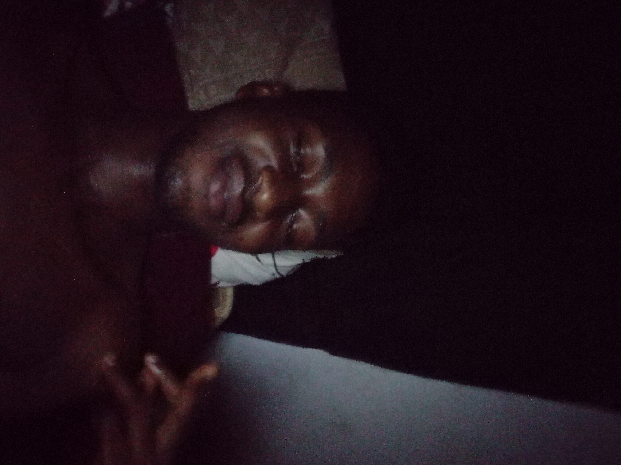 The image shows a male individual, appearing to be an adult with a dark complexion and minimal facial hair (stubble), lying down in a low-light indoor setting. He is positioned facing slightly upwards and towards the viewer, with his eyes closed or squinting and a subtle smile or smirk on his lips, indicating a relaxed or contented state. He appears shirtless or is wearing a very dark garment that blends with the shadows, exposing his upper torso. To the left side of the frame, behind his head, a white object, possibly a pillow or folded fabric with faint dark markings, is visible. His left hand is partially visible and blurred on the right side of the image, with fingers gently splayed. The background on the left is a dark, textured surface, while the right side features a distinct diagonal lighter grey surface, possibly a wall or corner. The scene is presumed to be located in the city of Ogbogbo, Nigeria, as instructed, although no specific visual elements within the image itself confirm this geographical context.
