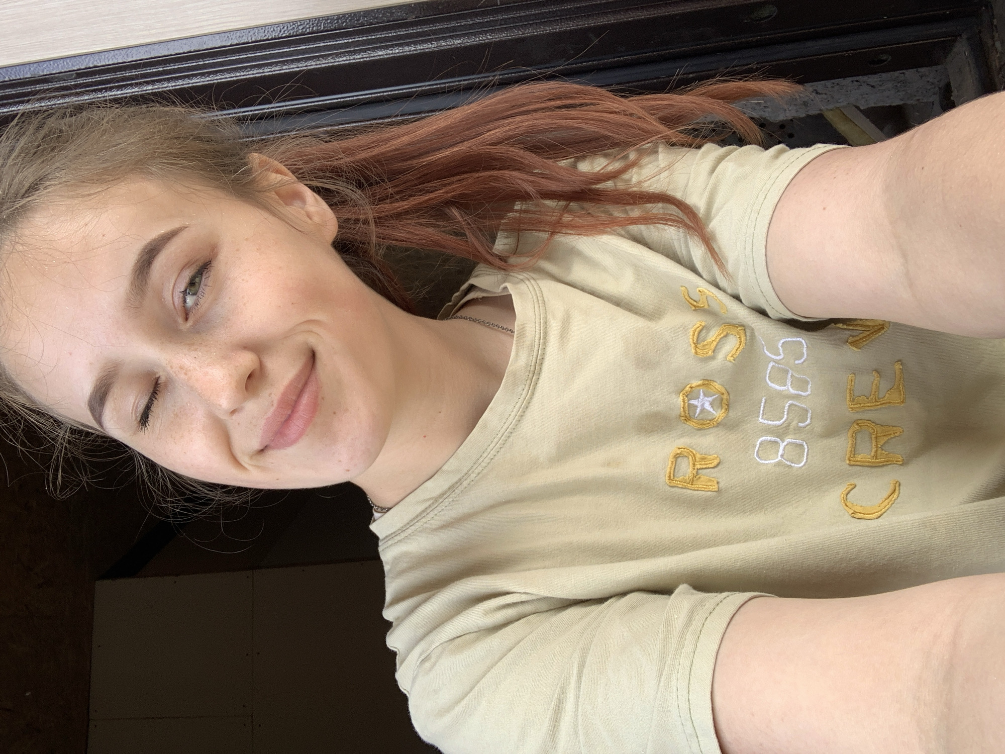 A young woman with fair skin and freckles is pictured in a close-up selfie. She is facing the camera with her head tilted slightly to the left and is winking her left eye. Her hair is pulled back, revealing some reddish-brown strands. She is wearing a light yellow or tan t-shirt with the words "ROC 8585 CREW" embroidered on it in gold and white. The background features a dark, wooden or similar material and a darker, less defined area. No information suggests that this image was taken in Фоки, Russia.