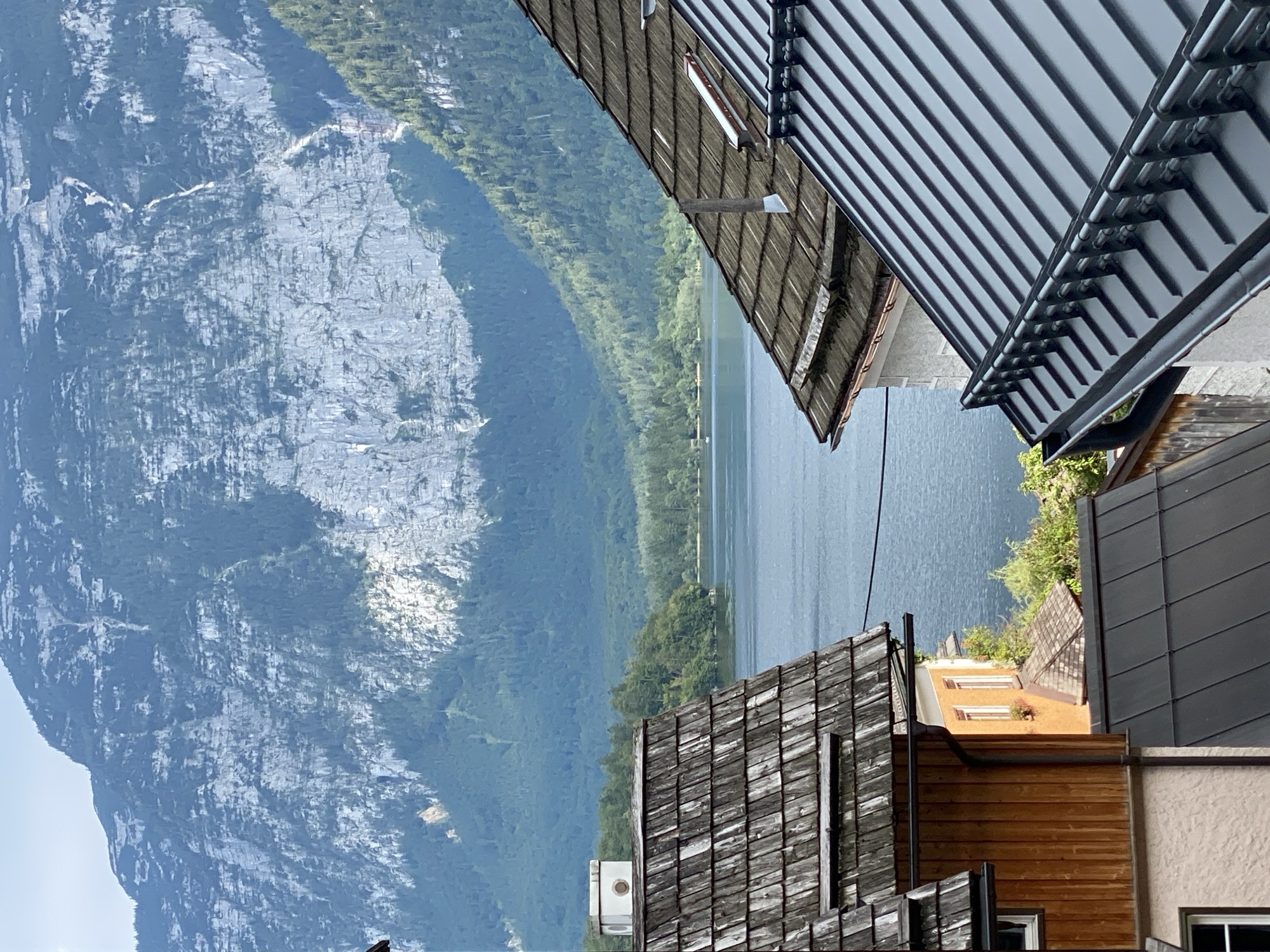 The image shows a view of Hallstatt, Austria, featuring traditional Austrian architecture and the stunning natural landscape. In the foreground, several rooftops are visible, with varying materials and styles. The roof on the left is made of weathered wooden shingles, showing signs of age. To its right, another roof with similar wooden shingles is partially obscured. Further to the right, a modern, dark gray metal roof with a prominent gutter system dominates the lower right portion of the frame.
In the middle ground, a serene lake reflects the sky and surrounding scenery. On the far side of the lake, lush green trees form a dense forest that extends up the slopes of the mountains.
The background is dominated by a majestic, rugged mountain range with a textured, rocky face. The mountain is partly covered in vegetation, with patches of bare rock and some visible water streaks or small waterfalls. The sky above is a pale blue with some wispy clouds. The overall impression is one of a picturesque village nestled within a grand natural setting.