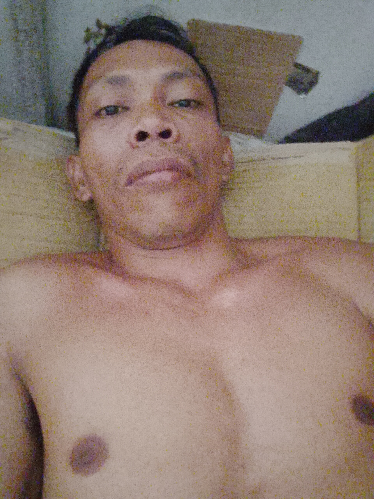 A shirtless man lies on a light-colored cushioned surface. He has dark hair and skin, and is facing the camera. His location is stated as Bukit Ketok, Indonesia, but the specific environment is not discernible beyond the presence of a cushioned surface and a partially visible item that could be furniture behind his head. There are no observable actions or interactions occurring.