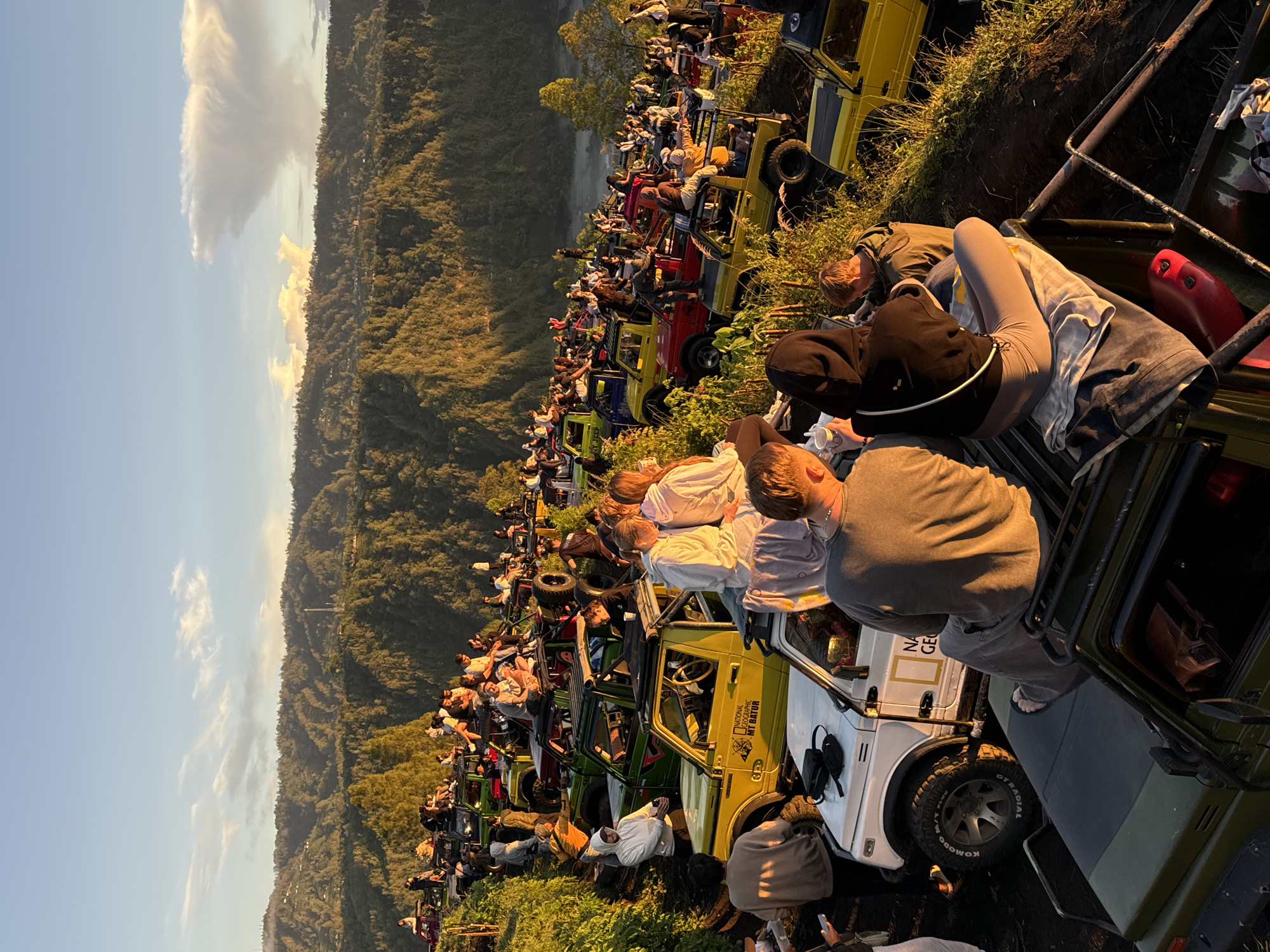 The image captures a bustling scene in Kintamani, Indonesia. Numerous open-top jeeps, painted in various colors like yellow, red, green, and white, are lined up along a roadside. People, dressed in casual clothing, are seated or standing in the jeeps, seemingly observing something in the distance. The backdrop features a landscape of mountains covered in dense greenery, set against a clear sky with a few scattered clouds. The overall atmosphere suggests an event where people have gathered to witness a scenic view or activity. The time of day appears to be early morning or late afternoon, judging by the soft, golden sunlight illuminating the scene.
