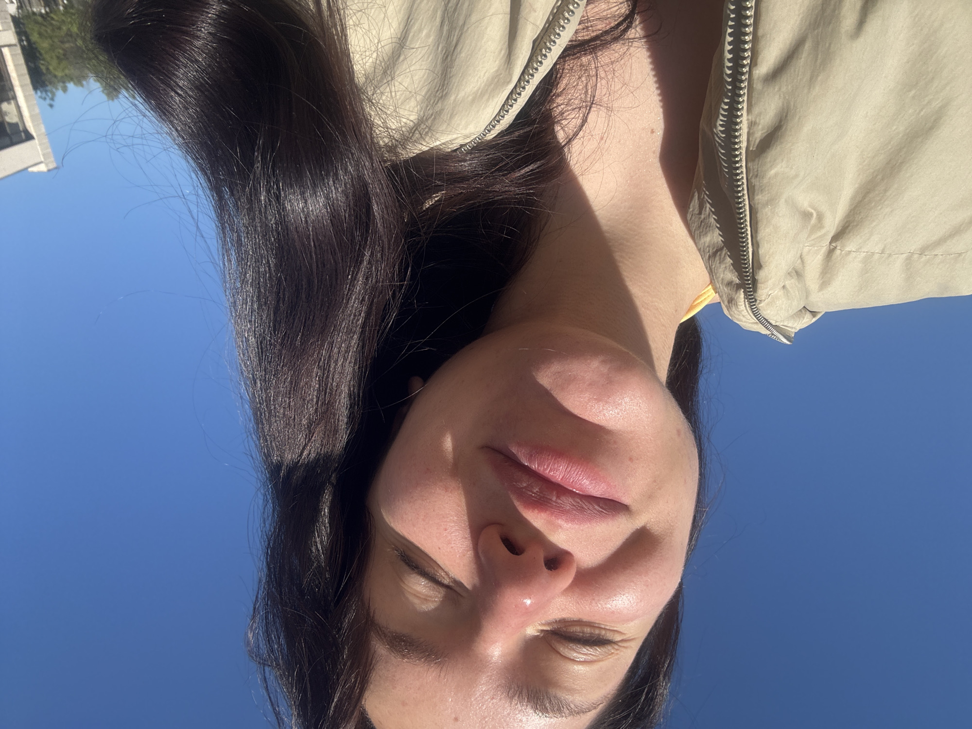 The image shows a woman's face from a low angle, positioned against a clear blue sky. She has dark hair, fair skin, and is wearing a beige jacket with a visible zipper. Her eyes are closed, and there is a slight shine on her nose. A portion of a building is visible in the top left corner of the image. The image appears to be taken outside on a sunny day. There is no information available in the image to confirm that the location is Blagoevgrad, Bulgaria.