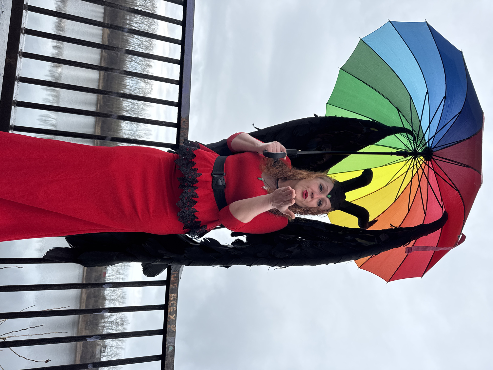 A woman in a red dress, black wings, and small black horns stands on a bridge, holding a rainbow-colored umbrella. She is blowing a kiss towards the camera. The bridge has a black metal railing with a body of water visible in the background. The sky is overcast. The location is Chaikovsky, Russia.
