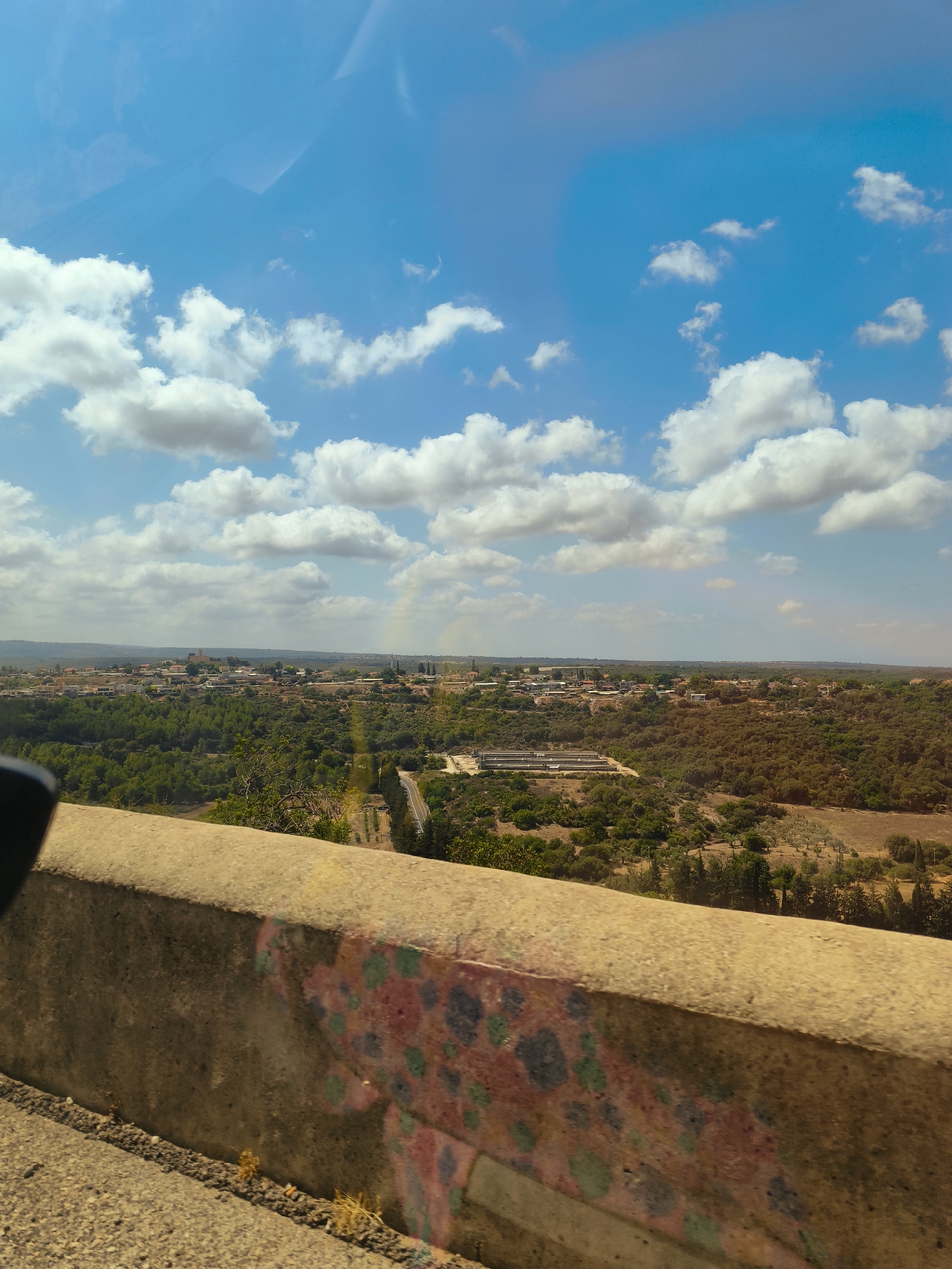 The image shows a landscape view of the city of Mate Asher Regional Council, Israel. In the foreground, there is a concrete barrier with colorful graffiti. Beyond the barrier, the scene unfolds with a rolling landscape covered in green trees and some arid terrain. A town or village is visible in the mid-ground, with scattered buildings and structures. The sky is a vibrant blue with scattered white clouds, and a faint rainbow is visible in the distance. The overall impression is a sunny day in a semi-arid region with a human settlement nestled within the natural environment.