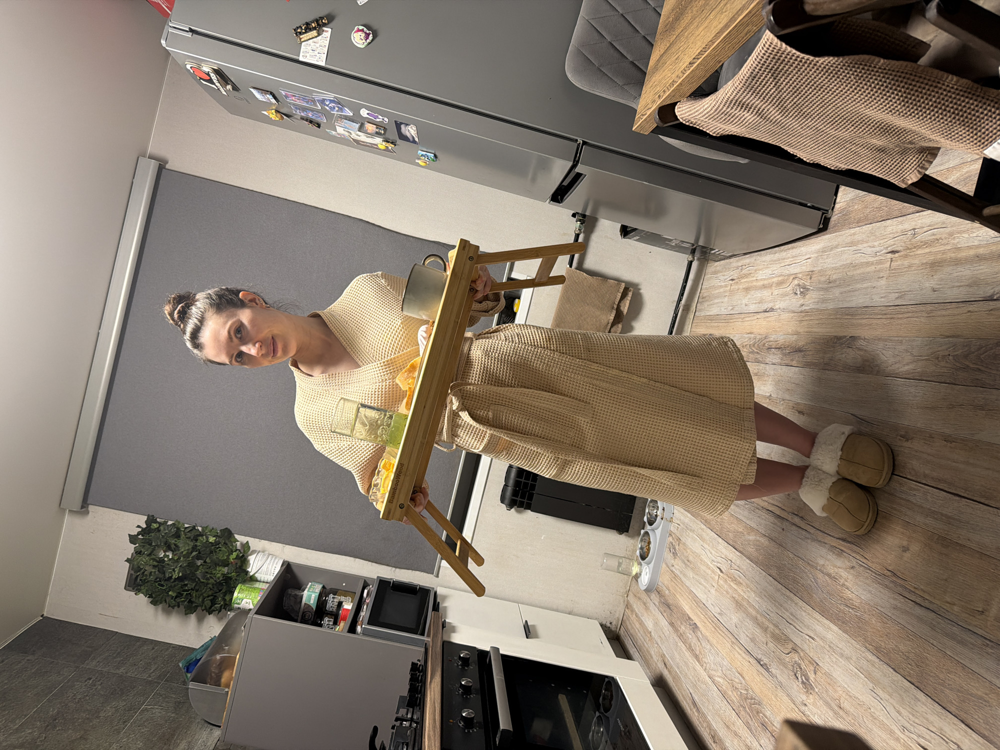 The image shows a woman in a tan waffle-patterned robe and slippers standing in what appears to be a kitchen. She holds a wooden breakfast tray containing a mug, juice, and other items. The kitchen features a gray refrigerator with magnets, a gray countertop, and light brown wood-look flooring. In the background, there is a blind and various kitchen appliances are visible, including an oven. The photo doesn't provide sufficient details to confirm if the location is Perm, Russia.