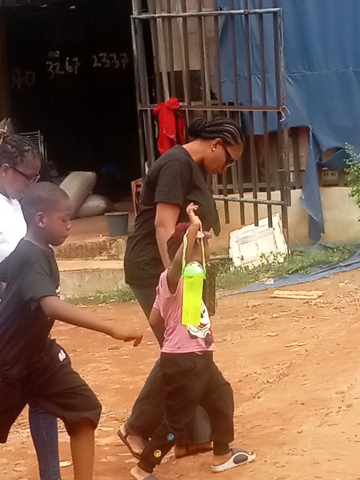 The image shows a group of people in Umuokpu, Nigeria. A woman with braids and sunglasses is standing near a child who is holding a green water bottle. To the left, a young boy in a black shirt and shorts is walking. Another woman is partially visible in the background. They appear to be outdoors on a dirt or sandy surface. Behind them is a building with a dark entrance and white numbers "10 3267 2337" painted on the wall. A metal gate is visible, with a red cloth hanging on it. In the background, there's a blue tarp covering an area.